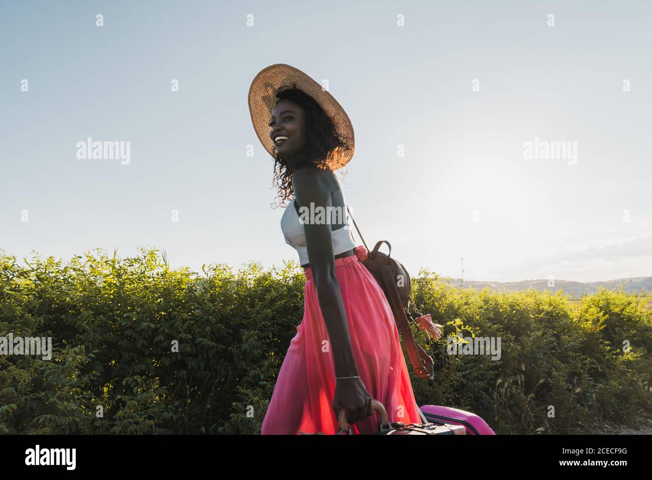 Trendige afroamerikanische Frau mit Hut, die Koffer trägt und im Sommer auf der Landstraße unterwegs ist Stockfoto