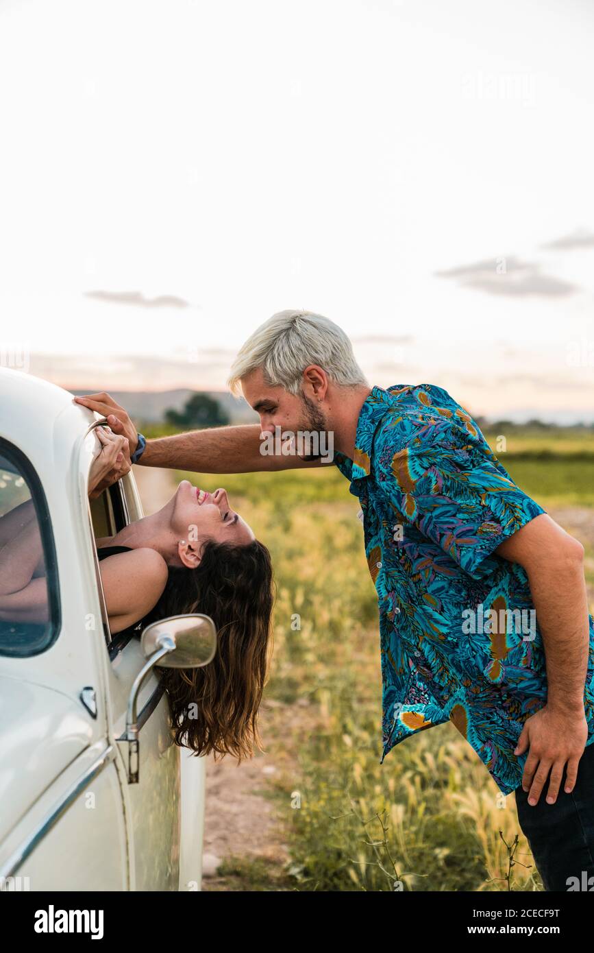 Seitenansicht einer Frau, die im Auto sitzt und sich mit dem Kopf nach außen lehnt und den Mann anschaut, der im Sommer glücklich darüber steht und lächelt Stockfoto