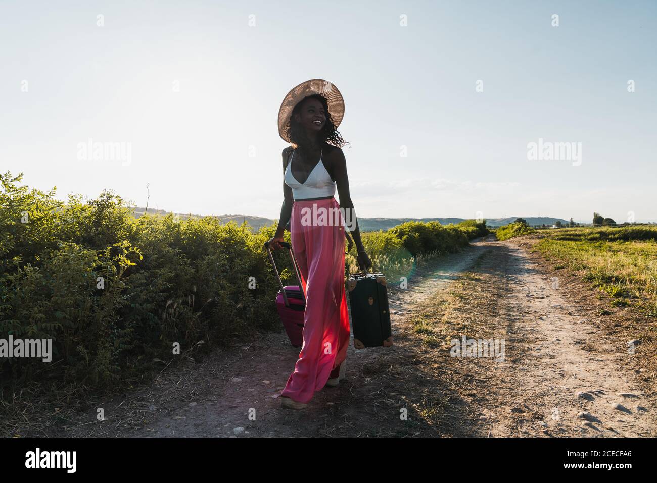 Trendige afroamerikanische Frau mit Hut, die Koffer trägt und im Sommer auf der Landstraße unterwegs ist Stockfoto