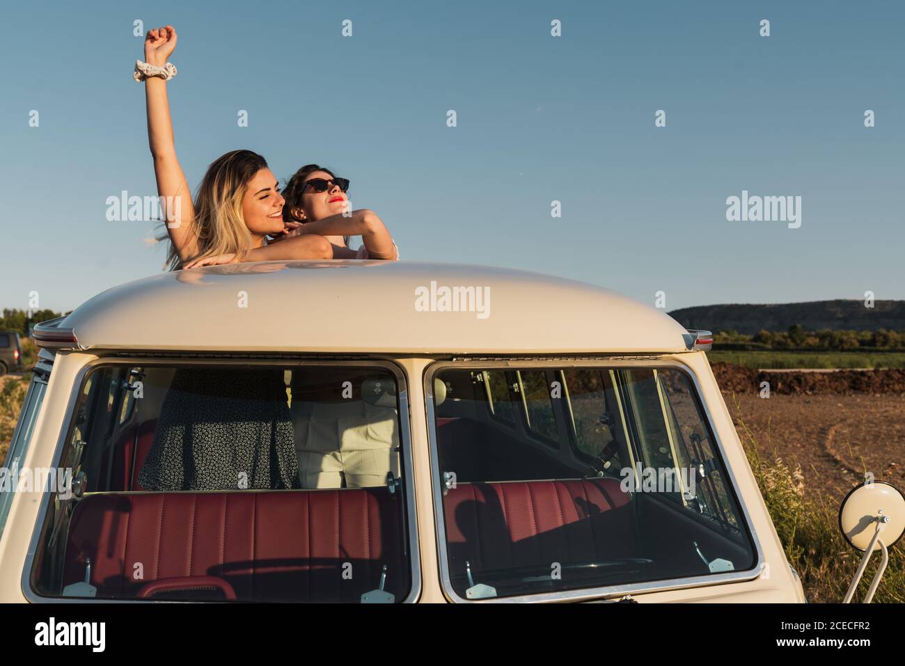 Zwei fröhliche junge Damen lachen und halten die Hände hoch, während Stehend in Luke im Dach des Vintage-Van in schön Landschaft an sonnigen Tag Stockfoto