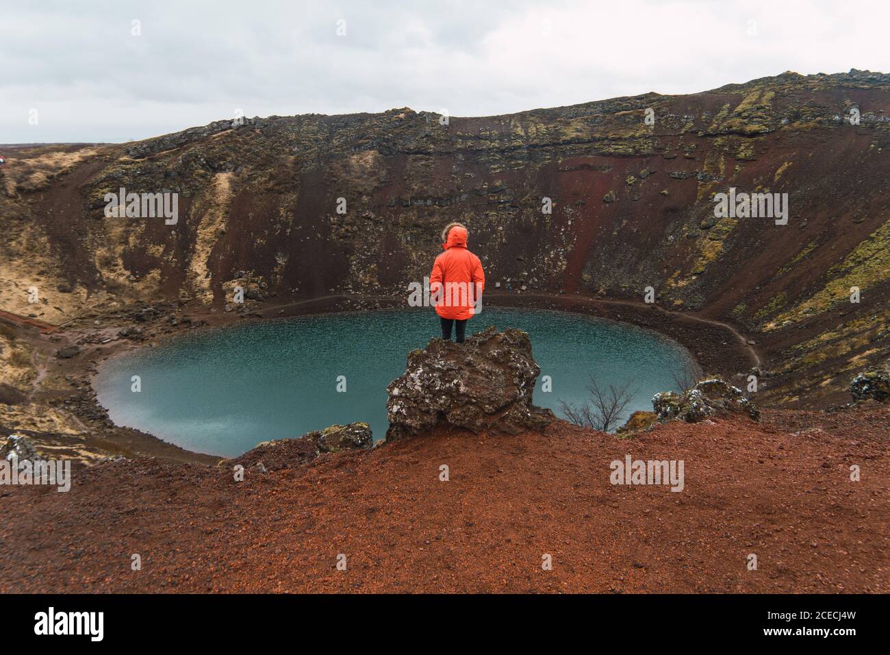 Rückansicht des Touristen in rotem Mantel stehend auf Felsen gegen kleinen blauen See im Becken des Berges in Island. Stockfoto