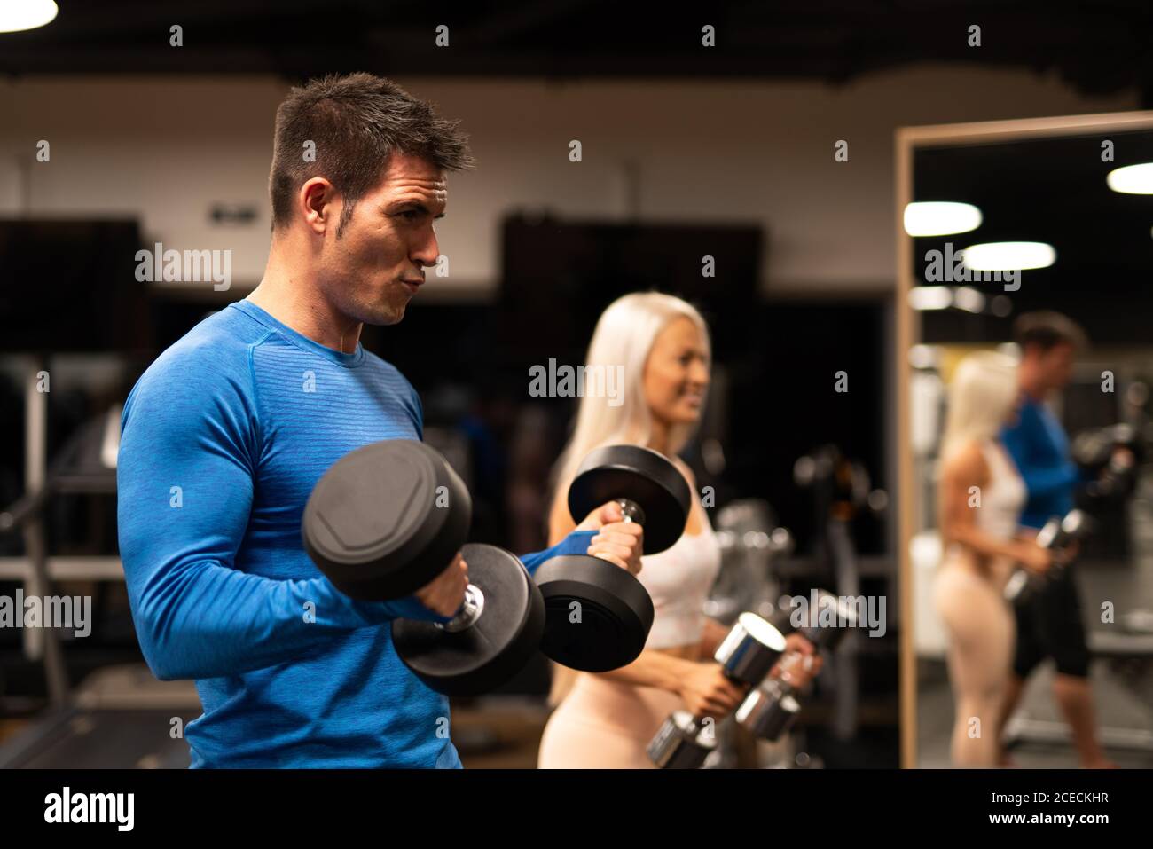 Seitenansicht eines hübschen Mannes mit blauem Pullover und einer attraktiven Frau in Sportkleidung, die im Fitnessstudio vor dem Spiegel steht und mit Hanteln trainiert Stockfoto