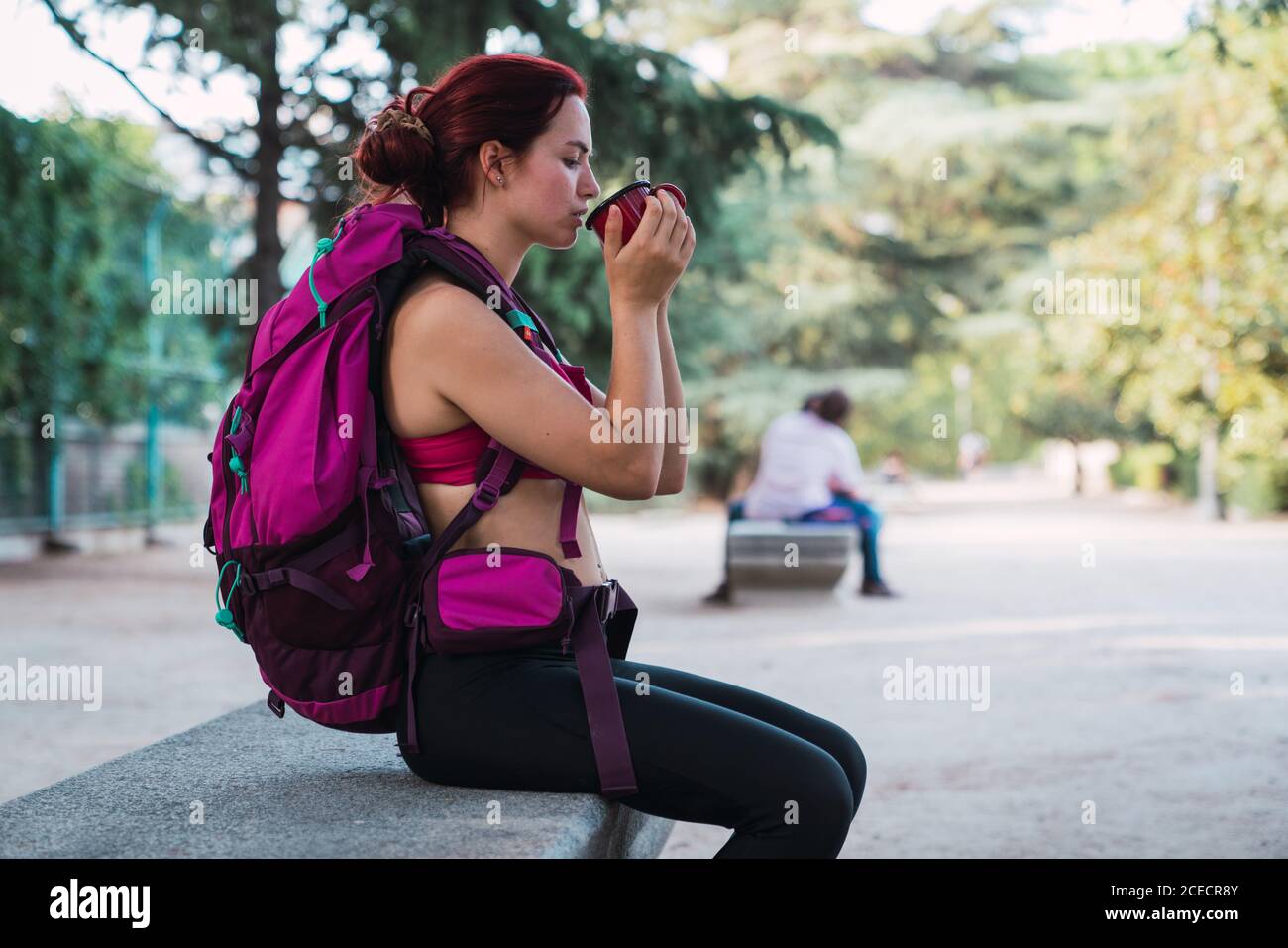 Seitenansicht einer schönen Frau in Sportkleidung mit großem rosafarbenem Rucksack, die auf einer Bank im Park sitzt und heißen Tee aus einer Metalltasse trinkt Stockfoto