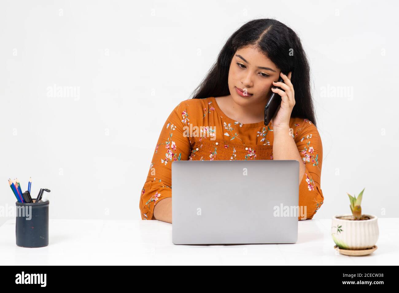 Hübsche junge Inderin, die vor weißem Hintergrund sitzt und ihren Laptop benutzt. Netter Teenager, der ihre Online-Schularbeit von zu Hause aus macht. Stockfoto