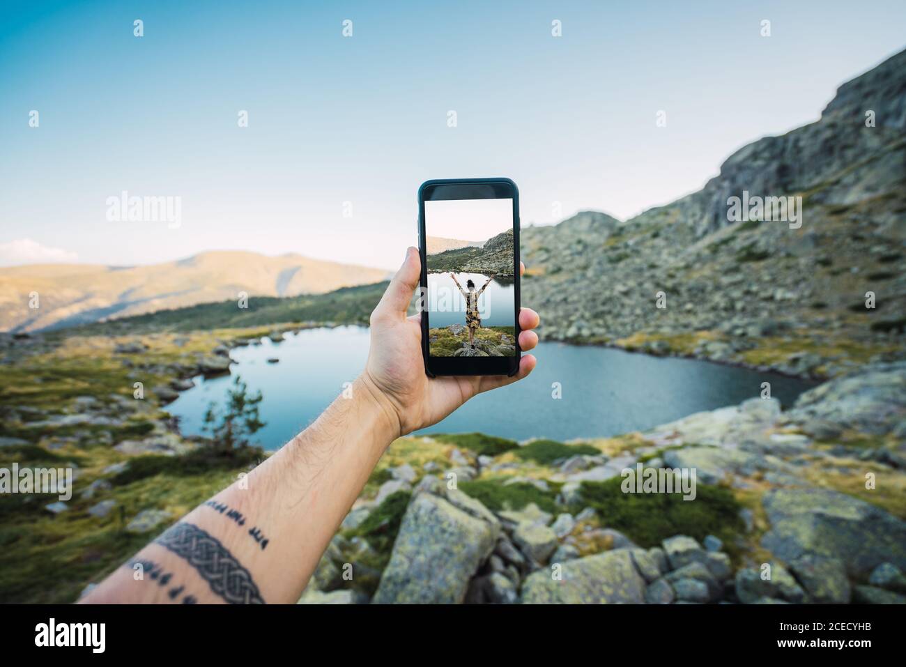 Crop tätowierte männliche Hand hält Smartphone mit Bild einer Frau auf dem Felsen stehen am Ufer des Bergsees Stockfoto