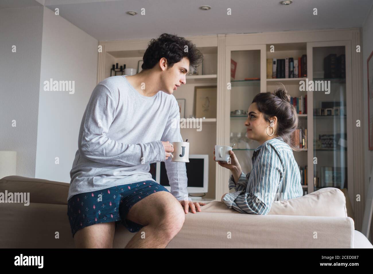 Glückliches junges Paar im Schlafanzug sitzt auf dem Sofa im Wohnzimmer mit Tassen. Stockfoto
