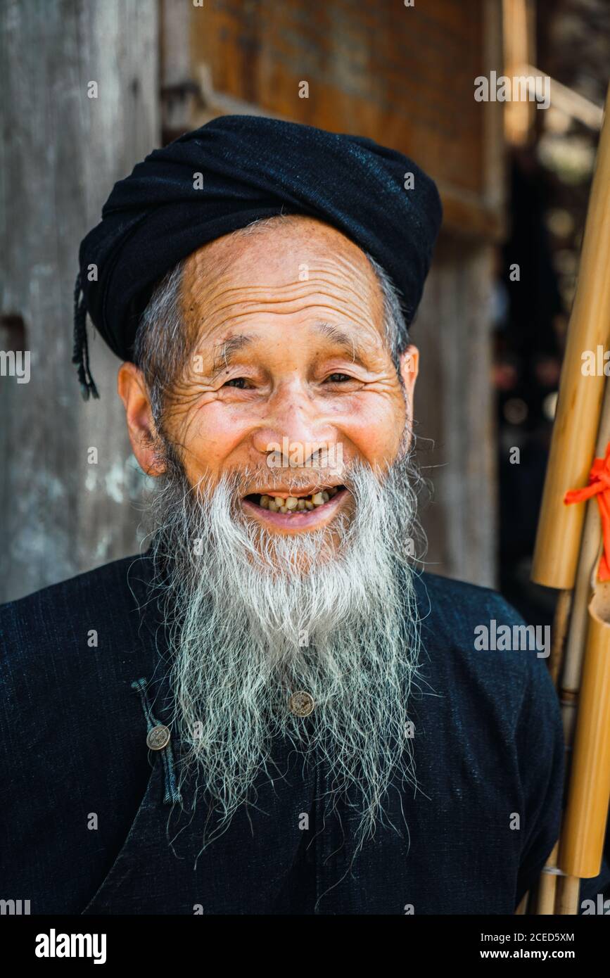 Fröhlicher alter Rüde mit langem grauen Bart in dunkler Kleidung Stehen mit Musikinstrument an der Wand auf schmalen chinesischen Dorf?Gasse Und mit anderen Menschen aus der Miao-Ethnie in die Kamera schauen Minderheit auf verschwommenem Hintergrund in der Provinz Guizhou Stockfoto