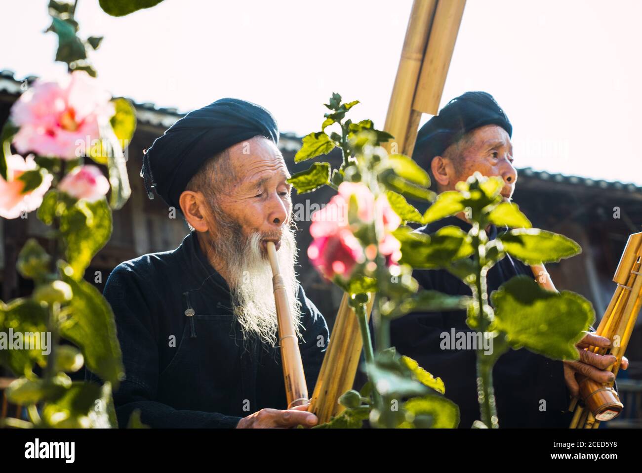 GUINZHOU, CHINA - 14. JUNI 2018: Ältere Männer in dunklen ethnischen Kostümen von Miao Menschen, die traditionelle Bambusflöten spielen, stehen am Sommertag im Freien beim Bauen mit hellen Blumen im Vordergrund in der chinesischen Provinz Guizhou Stockfoto
