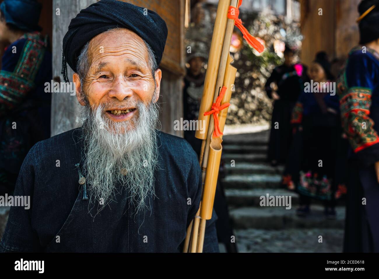 Fröhlicher alter Rüde mit langem grauen Bart in dunkler Kleidung Stehen mit Musikinstrument an der Wand auf schmalen chinesischen Dorfallee Und mit anderen Menschen aus der Miao-Ethnie in die Kamera schauen Minderheit auf verschwommenem Hintergrund in der Provinz Guizhou Stockfoto
