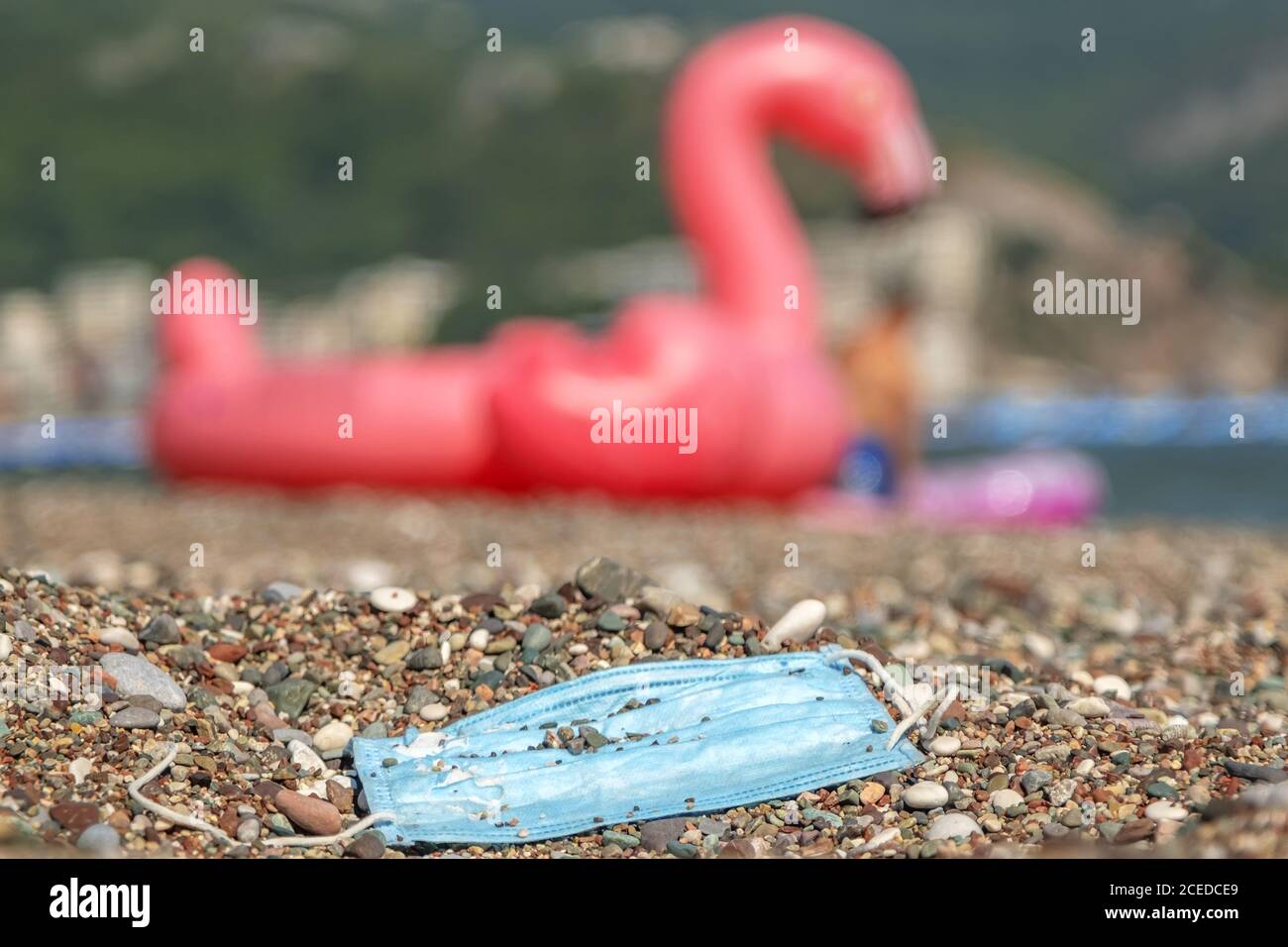 Ausrangierte gebrauchte Gesichtsmaske liegt an einem Kiesstrand, im Hintergrund ein aufblasbarer rosa Flamingo an der Küste. Stockfoto