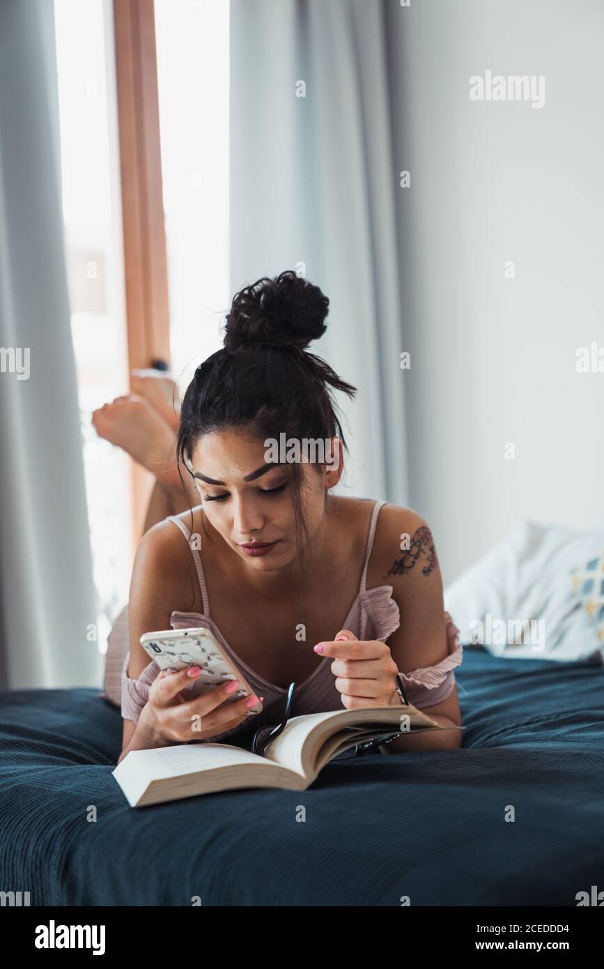 Hübsche Brünette entspannend zu Hause liegend auf dem Bett mit Buch Geöffnet und mit Smartphone in der Freizeit Stockfoto