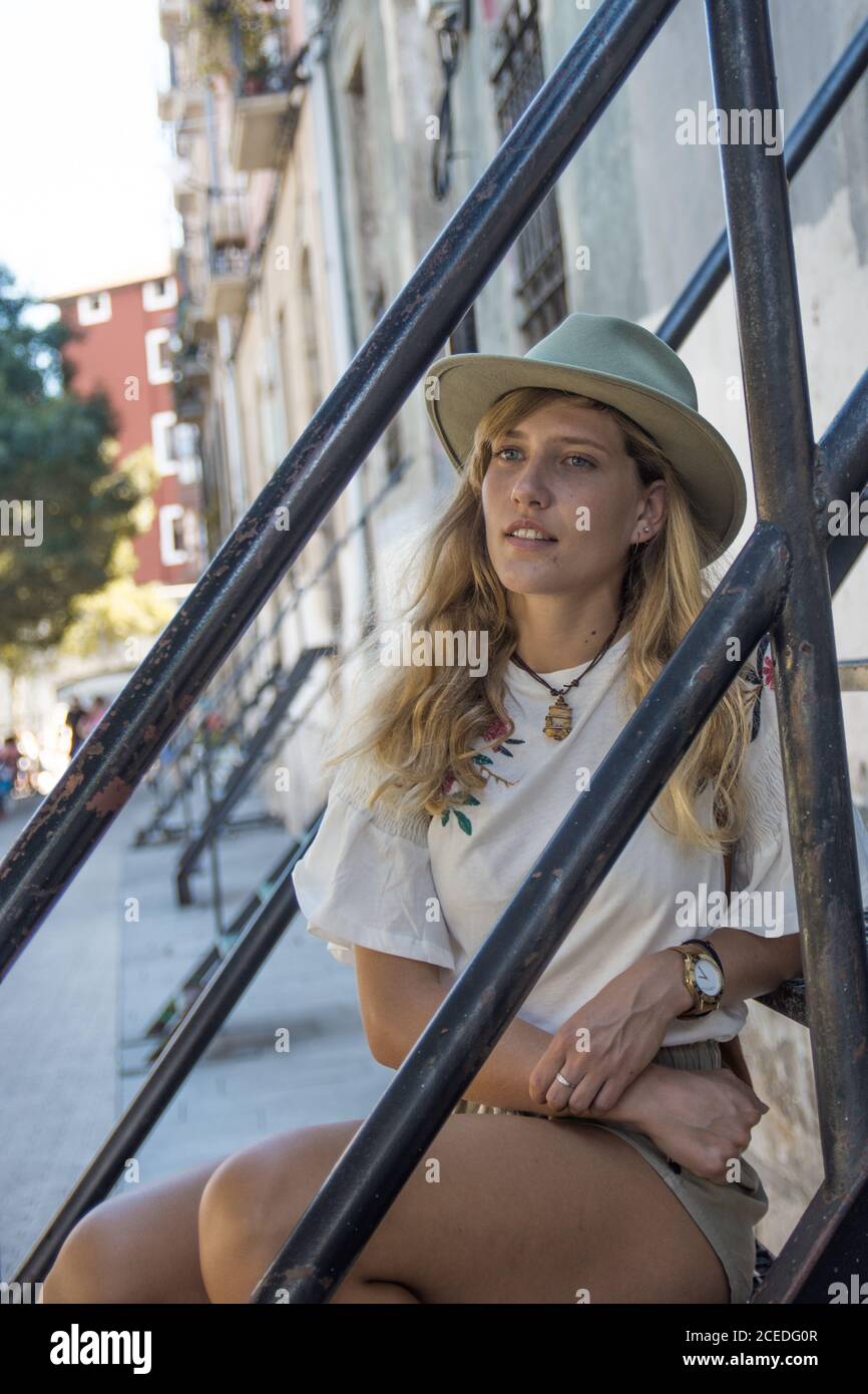 Junge blonde Frau im Hut und Sommer-Outfit auf den Stufen draußen sitzen und in Tagträumen wegschauen Stockfoto