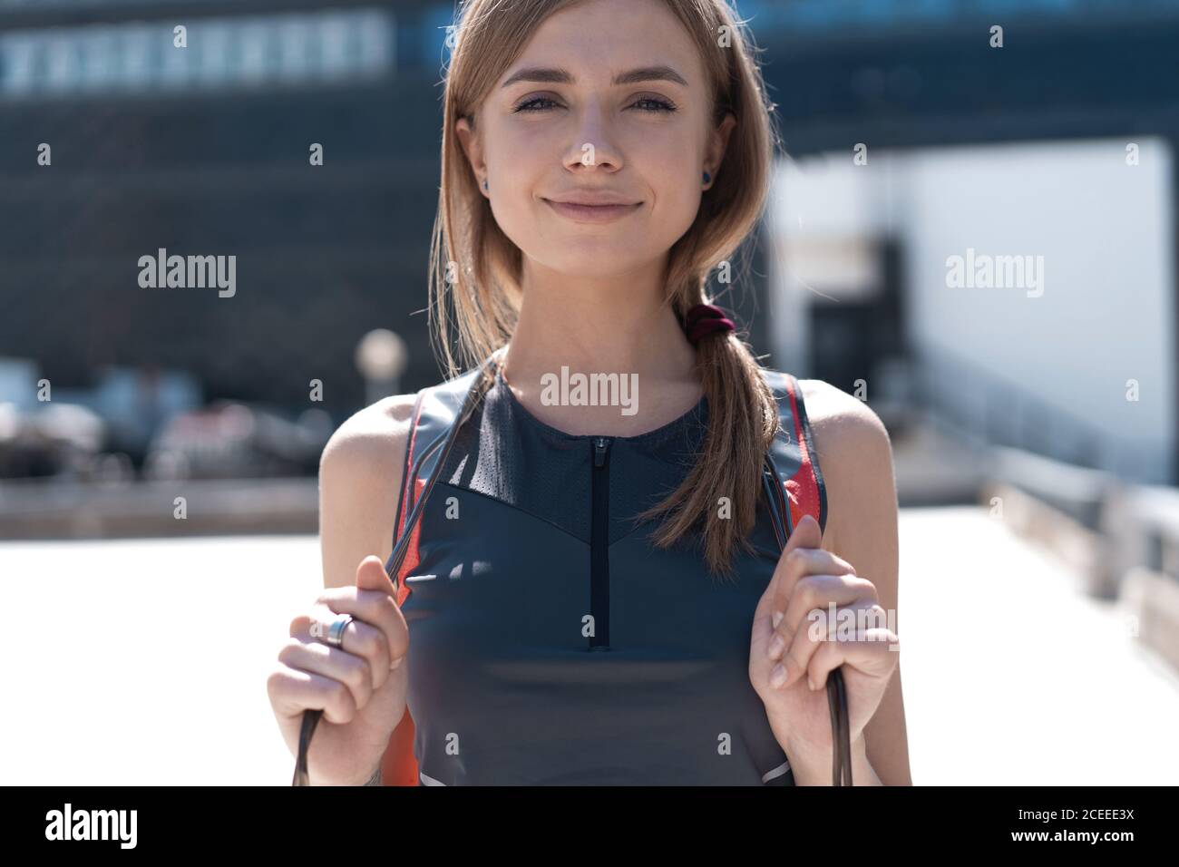 Glückliche junge Sportfrau im stylischen Trainingsoutfit mit Springseil auf ihren Schultern lächelt, draußen stehend Stockfoto
