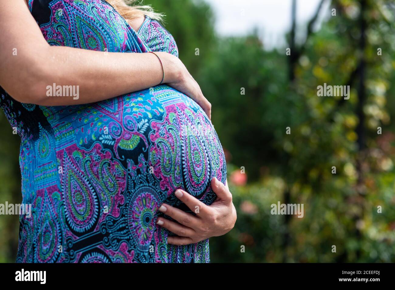 Seitenansicht Ernte schwangere Dame im Kleid hält Bauch in der Nähe Grüne Bäume auf verschwommenem Hintergrund Stockfoto