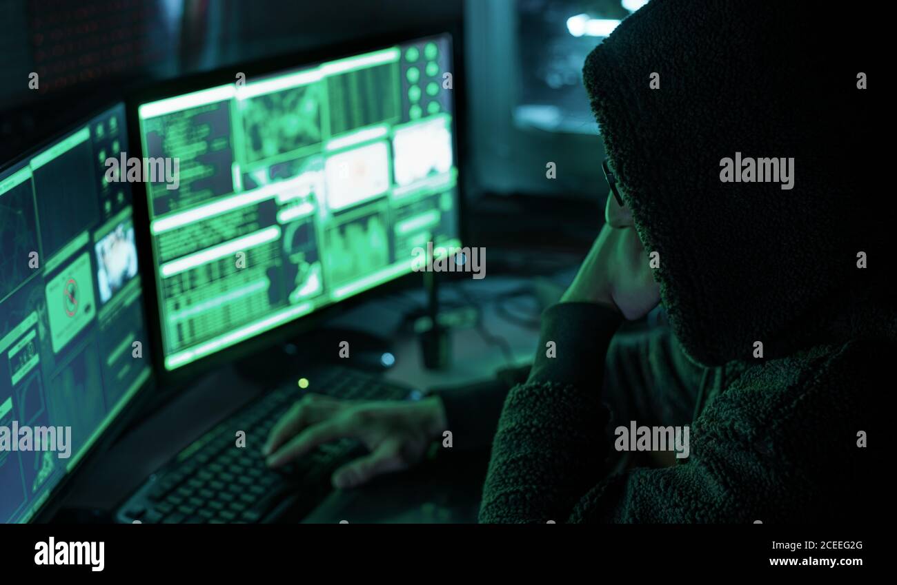 Gefährlicher Hacker mit Kapuze an seinem Versteck, der eine dunkle Atmosphäre, mehrere Displays hat Stockfoto