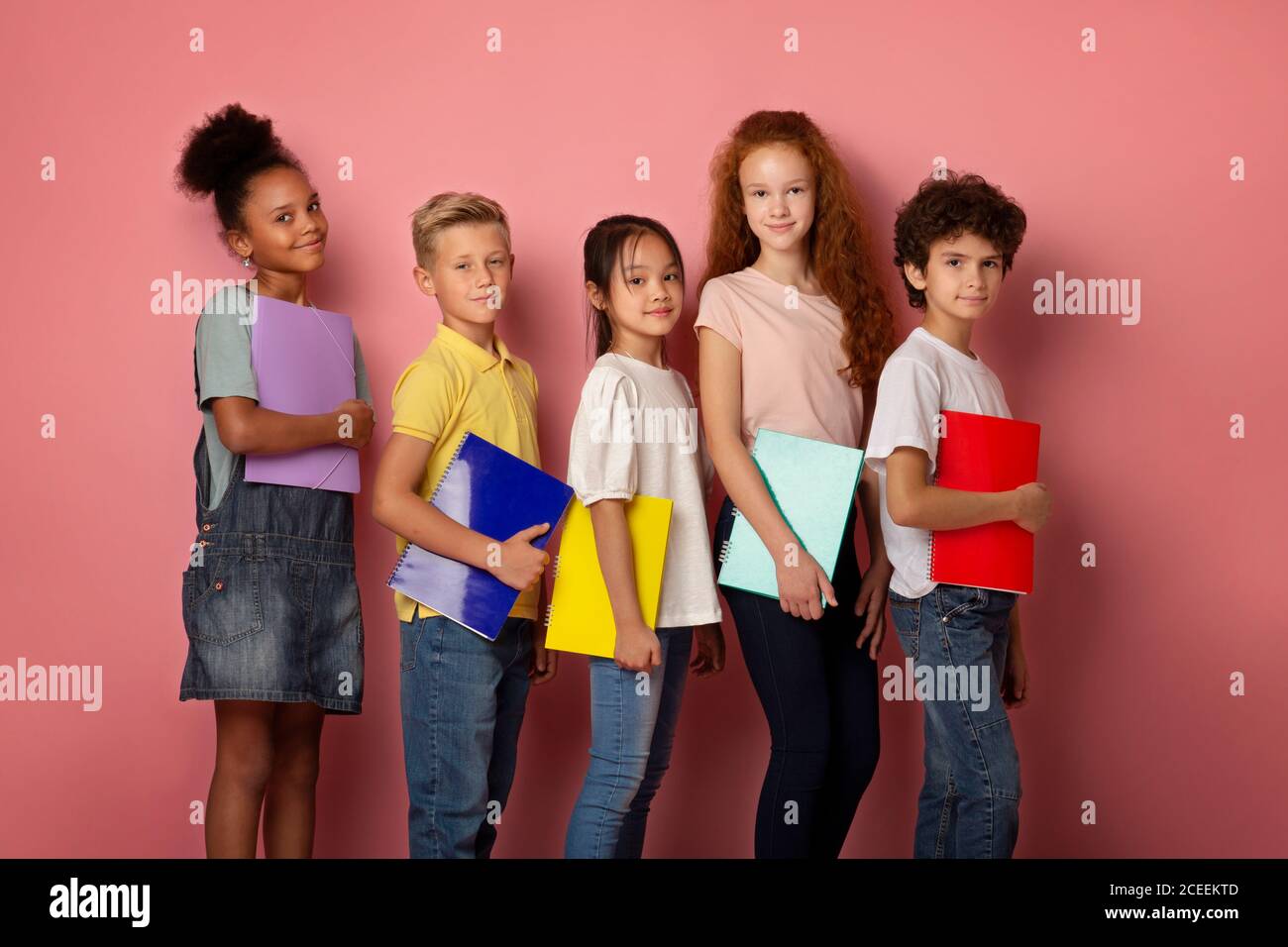 Fröhliche multiethnische Schüler stehen mit Notizbüchern in den Händen Rosa Hintergrund Stockfoto