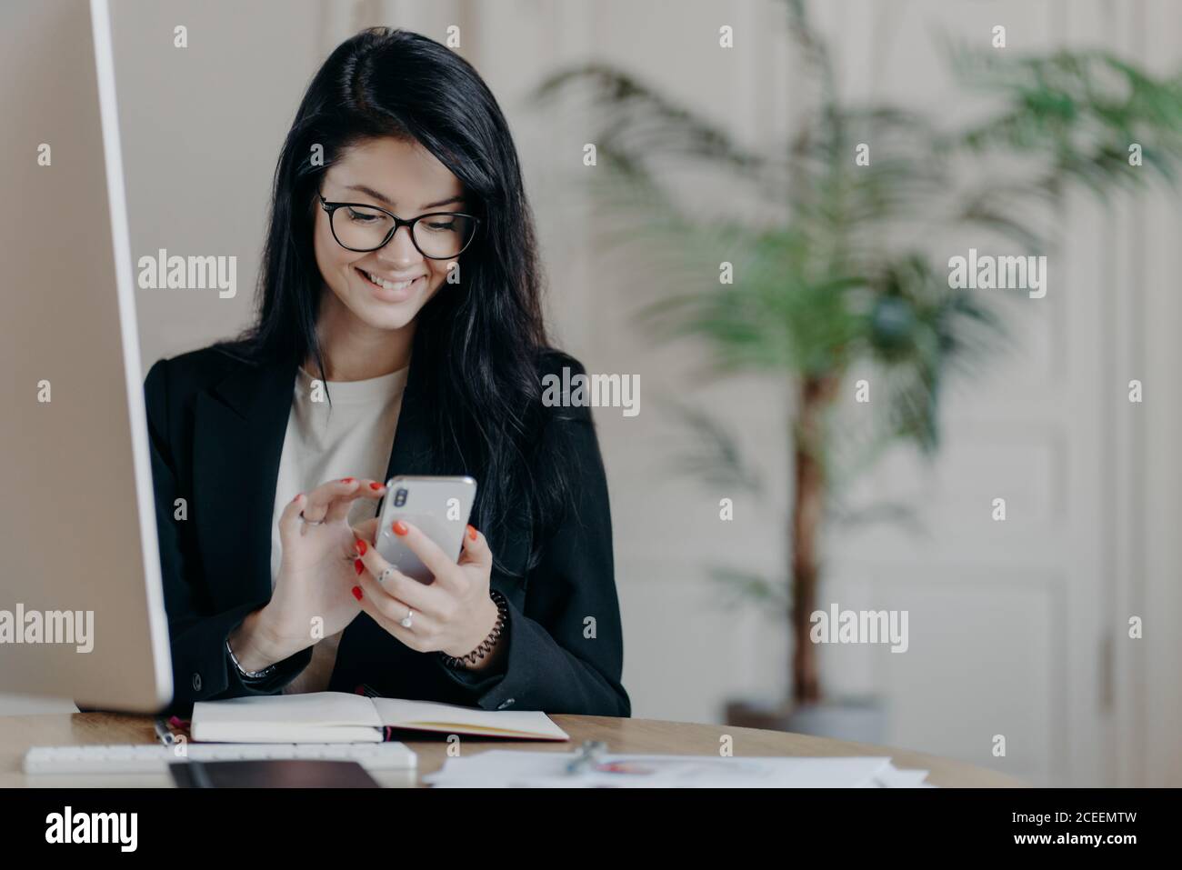 Schöne lächelnde Freiberuflerin arbeitet aus der Ferne, konzentriert in Smartphone, sendet Feedback auf empfangene Nachricht, sitzt im Coworking Space, gekleidet f Stockfoto