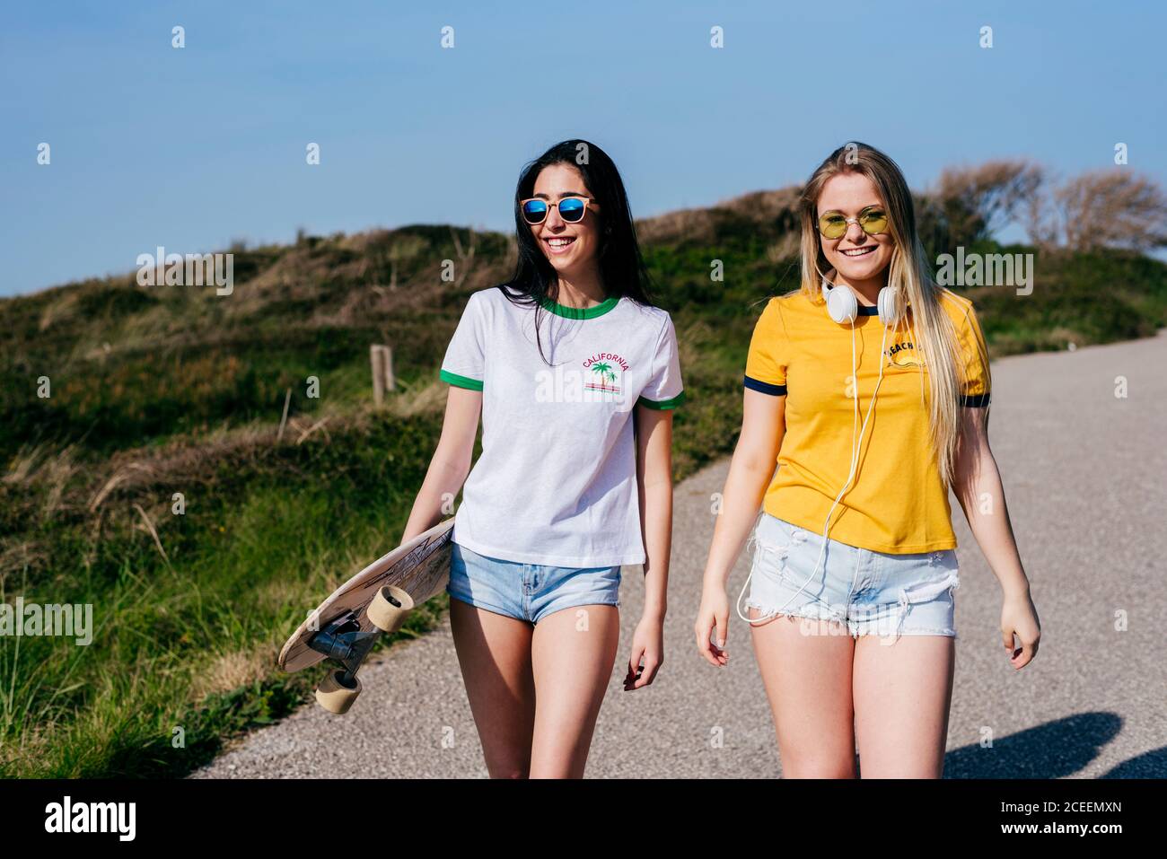 Hübsches junges Mädchen in Sommershorts zu Fuß auf ländlicher Straße mit langem Brett in strahlendem Sonnenschein und lächelnd. Stockfoto