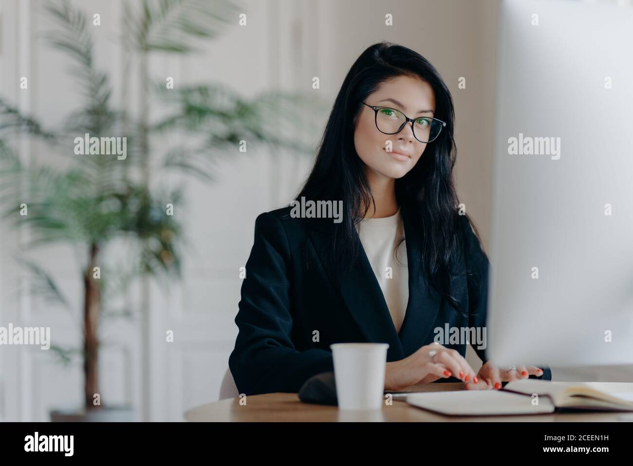 Seriöse erfolgreiche Büroarbeiterin in formeller Kleidung, trägt Brillen, beschäftigt mit Online-Arbeit, Posen am Desktop, arbeitet am Computer, liest Geschäft Stockfoto
