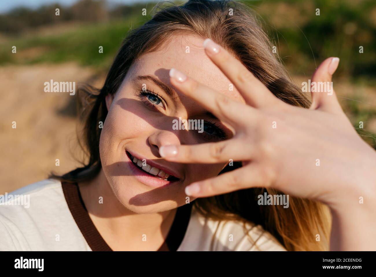 Liebenswert hübsches Mädchen in Sonnenschein macht Hand mit Handfläche auf Gesicht lächelt bei der Kamera im Sonnenlicht. Stockfoto