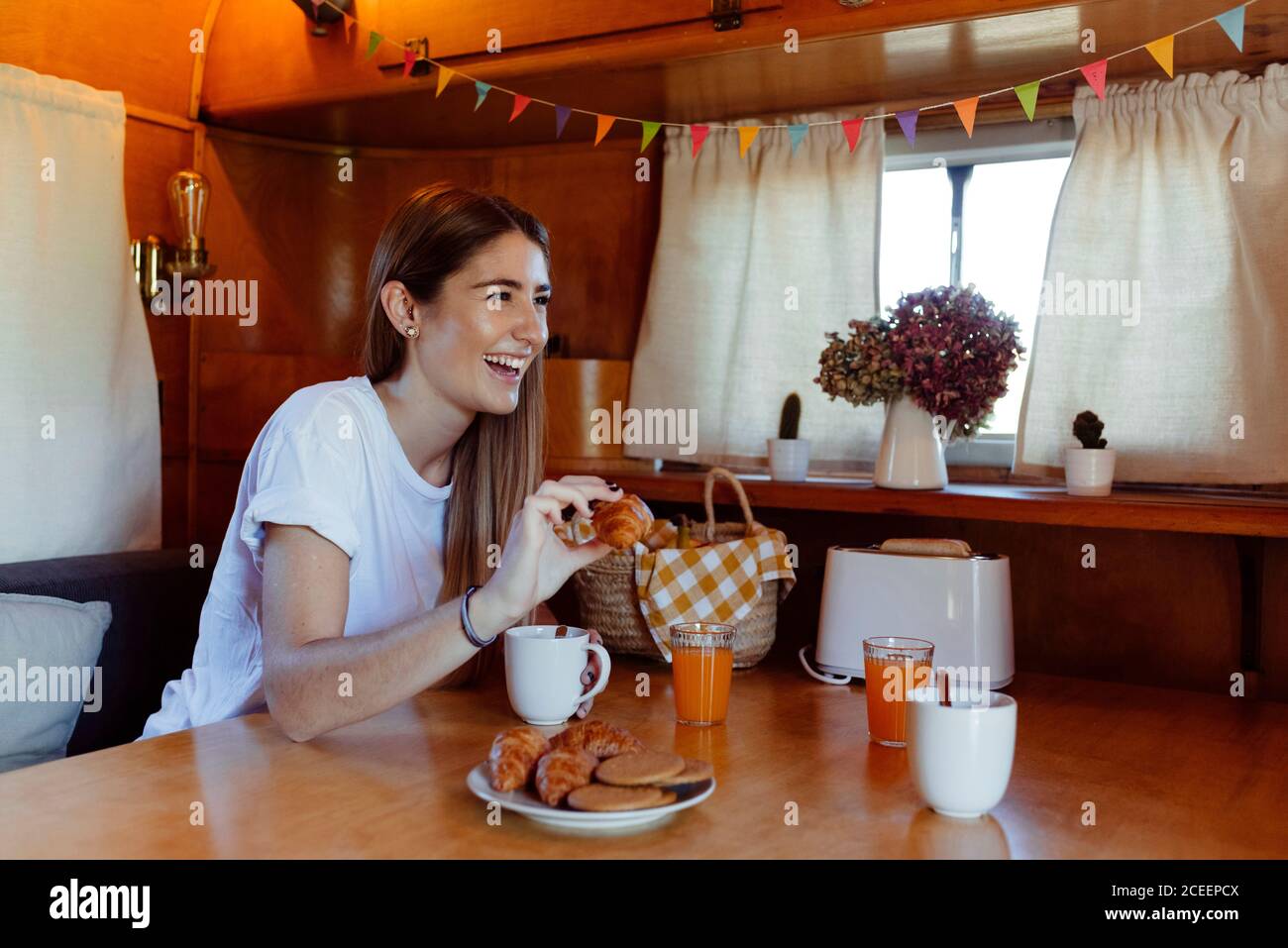 Reizende junge Frau, die ein kleines Brötchen hält und lacht, während sie im gemütlichen Van frühstückt Stockfoto