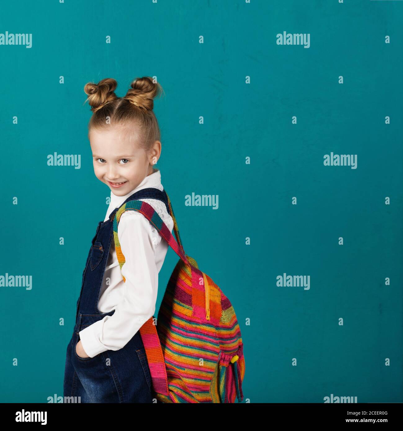 Schönes lächelndes kleines Mädchen mit großem Strickrucksack, der gegen die blaue Wand steht. Blick auf die Kamera. Schulkonzept. Zurück zur Schule Stockfoto