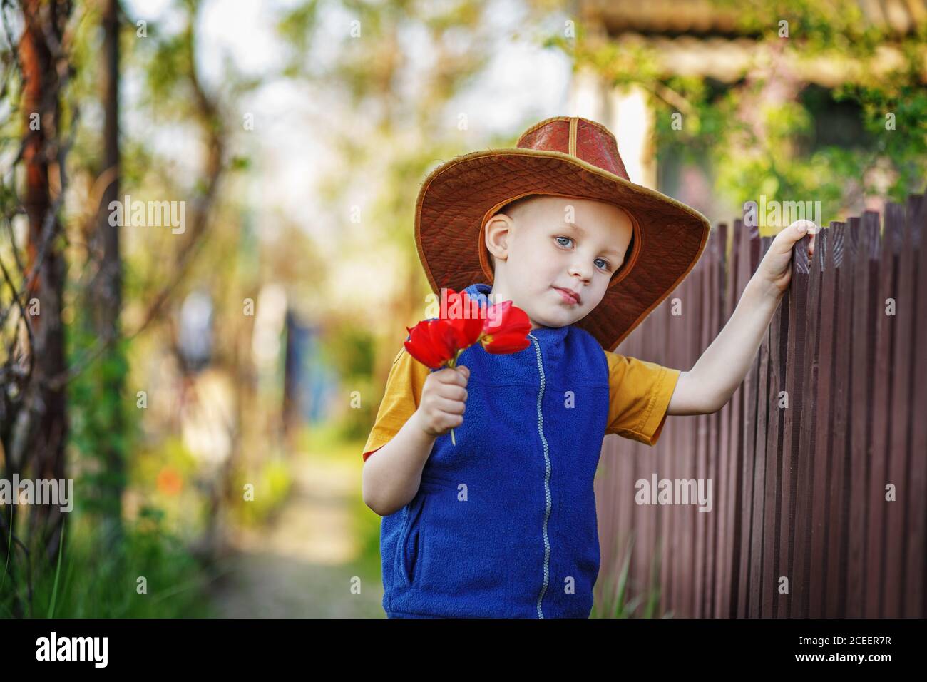 Porträt eines kleinen Jungen, der in einem großen Hut steht Mit einem Strauß Tulpen am Holzzaun in Die Landschaft Stockfoto