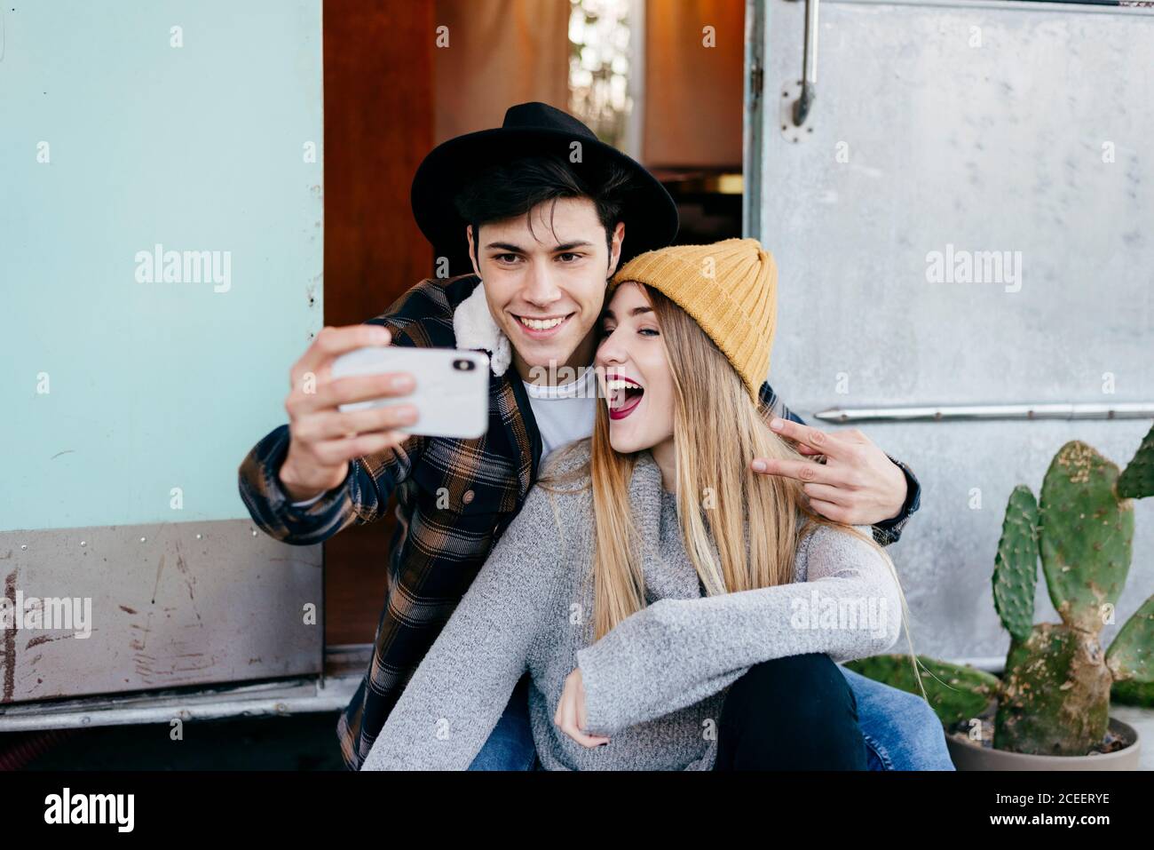 Hübscher junger Mann und schöne Frau, die lächeln und für Selfie posieren, während sie in der Nähe eines verwitterten Wohnwagens sitzen Stockfoto