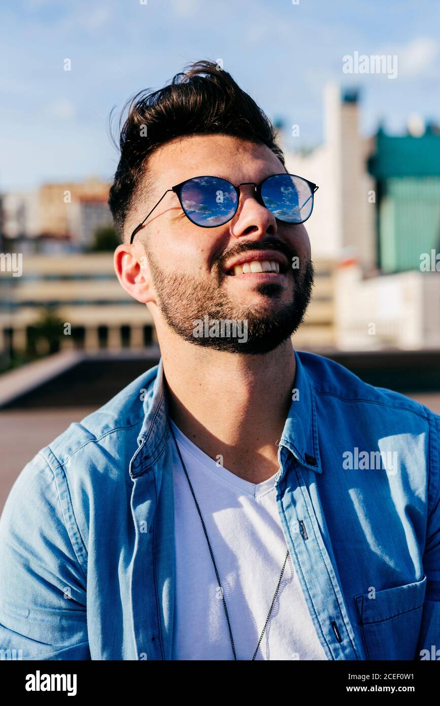 Schöner stilvoller Kerl mit Bart und moderner Frisur trägt Sonnenbrille und Denim-Shirt lächelt weg draußen. Stockfoto