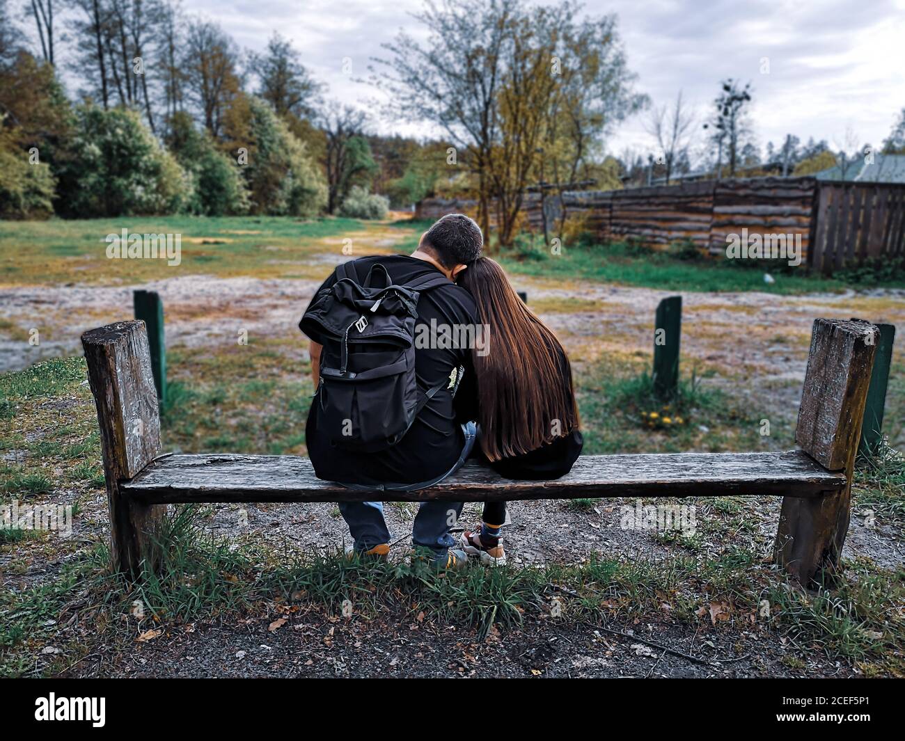 Rückansicht des Vaters mit Rucksack und der Tochter mit langen lockeren Haaren, die auf einer Bank mit dem Kopf auf den Schultern des anderen auf dem Land sitzt. Stockfoto