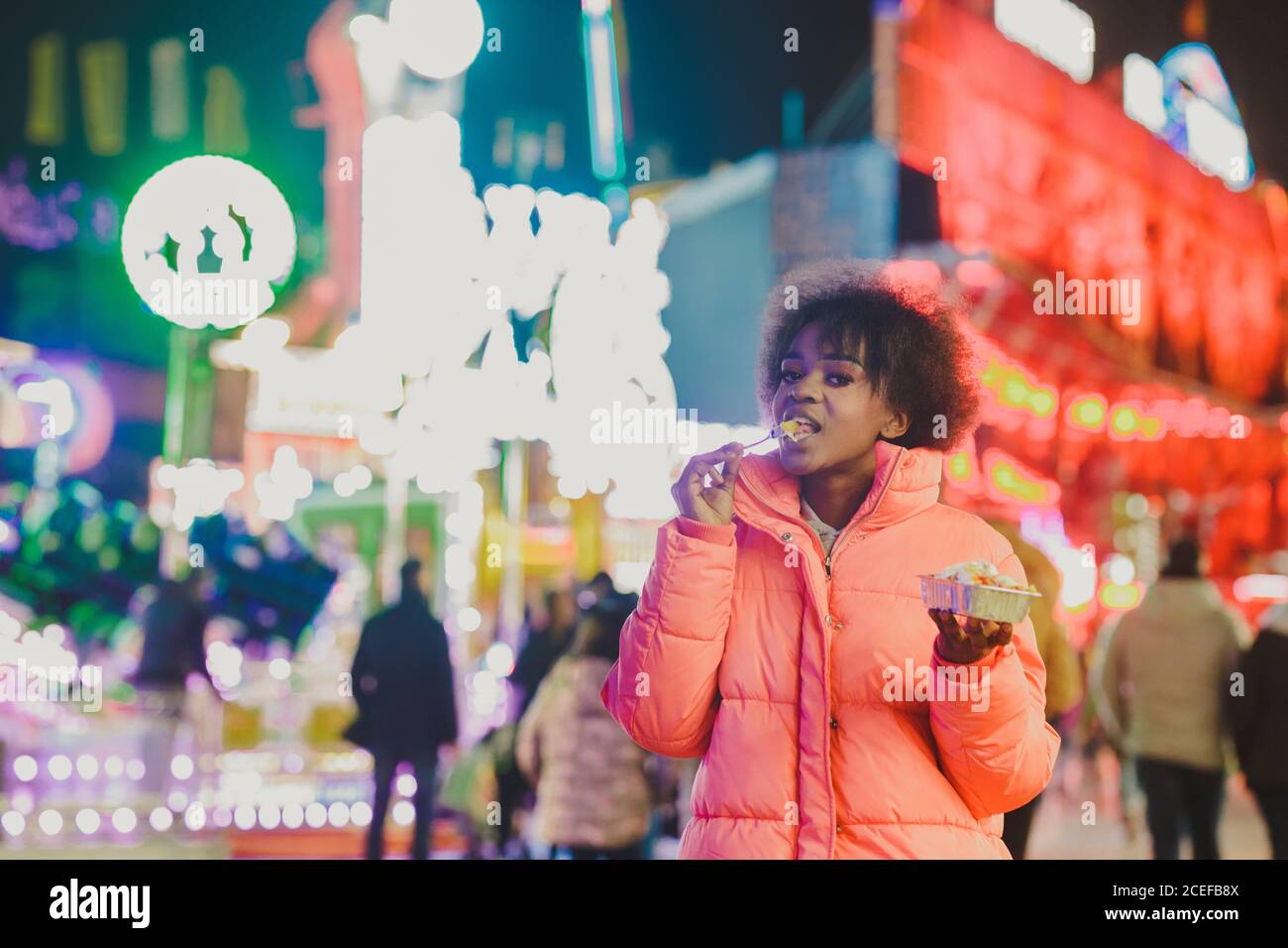 Hübsche afroamerikanische Dame in trendigen Kleidern essen leckeren Snack Und die Kamera betrachten, während sie auf unscharfem Hintergrund von steht Beleuchtetes Messegelände bei Nacht Stockfoto