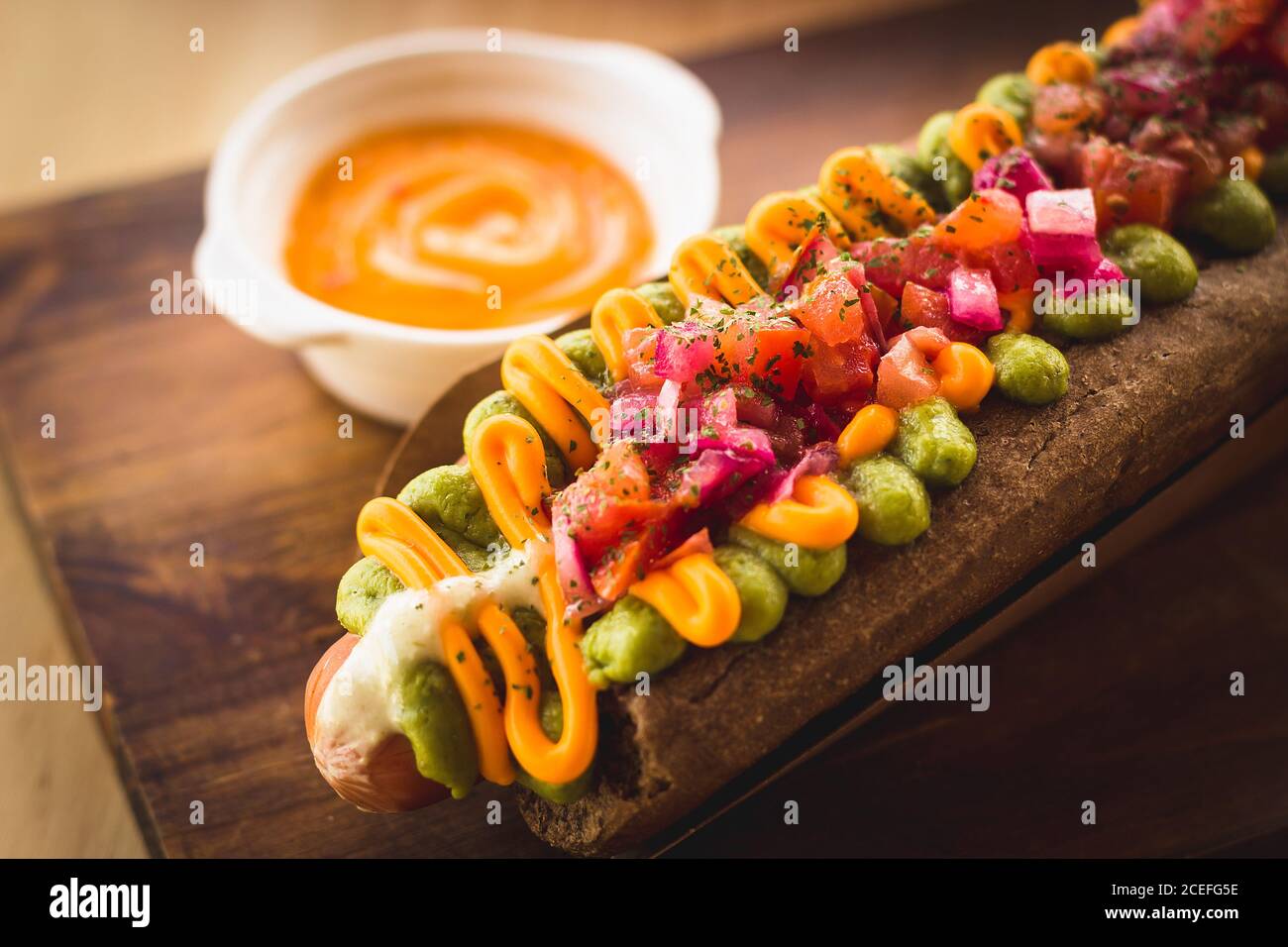 Nahaufnahme des servierten Hot Dogs mit schwarzem Brot Brötchen mit verschiedenen Saucen und Zusatzstoffen auf dem Teller serviert gekrönt. Stockfoto