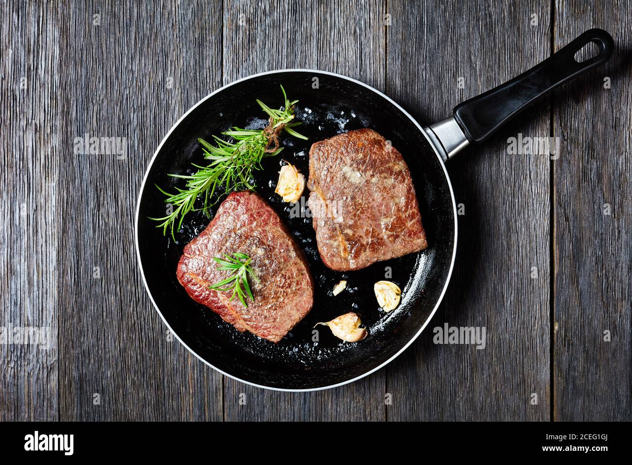 Gebratene Rindersteaks mit Rosmarinstrauß, Knoblauch und Salz auf einer Pfanne auf einem Holztisch, horizontale Ansicht von oben, flach liegend, Freiraum Stockfoto