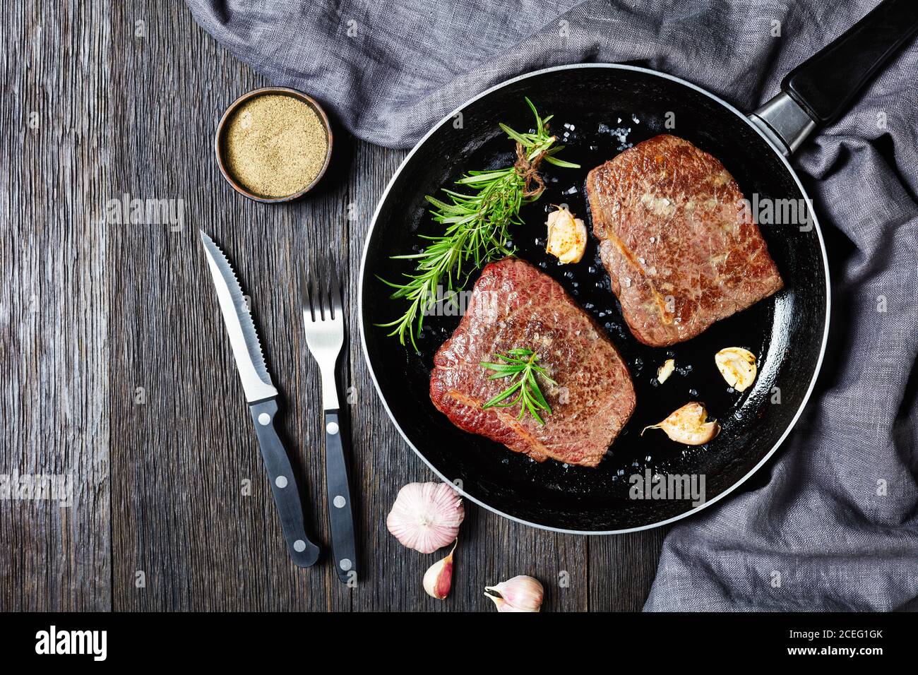 Gebratene Rindersteaks mit Rosmarinstrauß, Knoblauch und Salz auf einer Pfanne auf einem Holztisch mit Gabel und Messer, horizontale Ansicht von oben, flach liegend, fre Stockfoto
