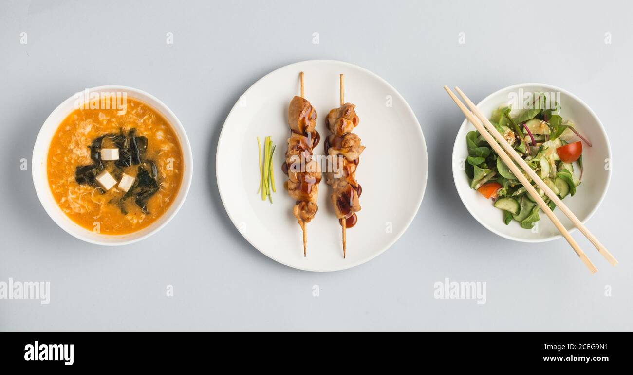 Schöne Schüsseln mit Miso-Suppe und frischem Gemüse Salat und Teller mit Sushi-Brötchen und Holzstäbchen liegen Auf weißem Hintergrund von oben Stockfoto