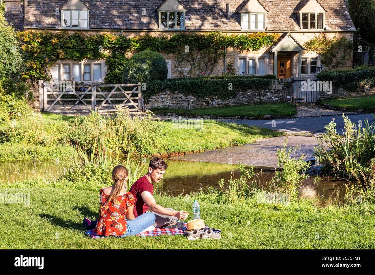 Ein junges Paar, das die Abendsonne neben der furt über dem River Eye im Cotswold Village of Upper Slaughter, Gloucestershire UK, genießt Stockfoto