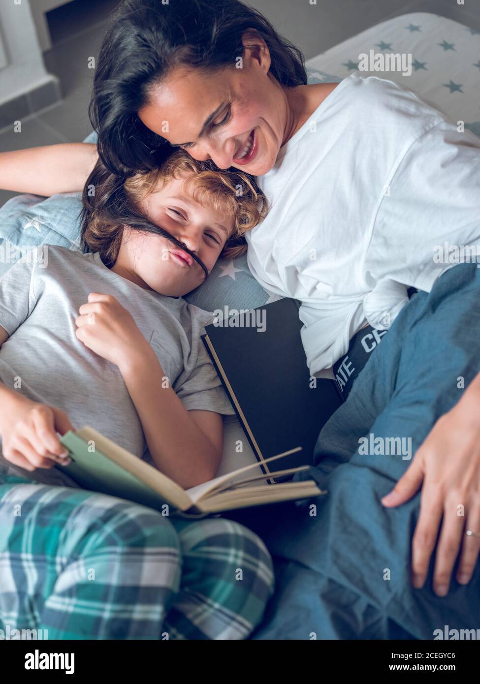 Netter Junge, der Spaß hat, während er auf dem Bett liegt und Interessantes Buch mit Hilfe der lächelnden Mutter liest. Stockfoto