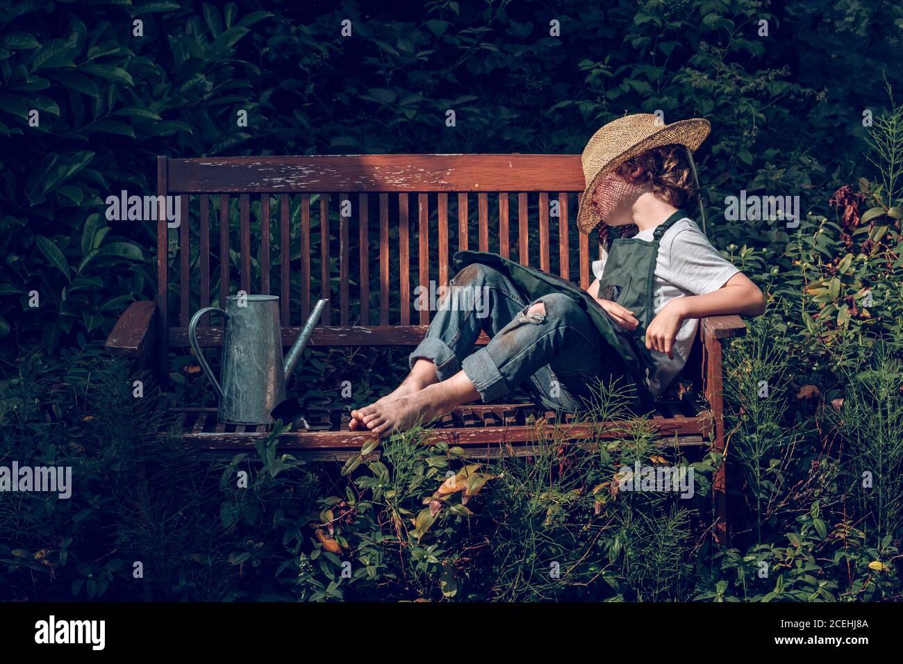 Kleiner Junge in Denim und Strohhut, der ein Nickerchen macht Holzbank nach der Arbeit im grünen Sommergarten Stockfoto
