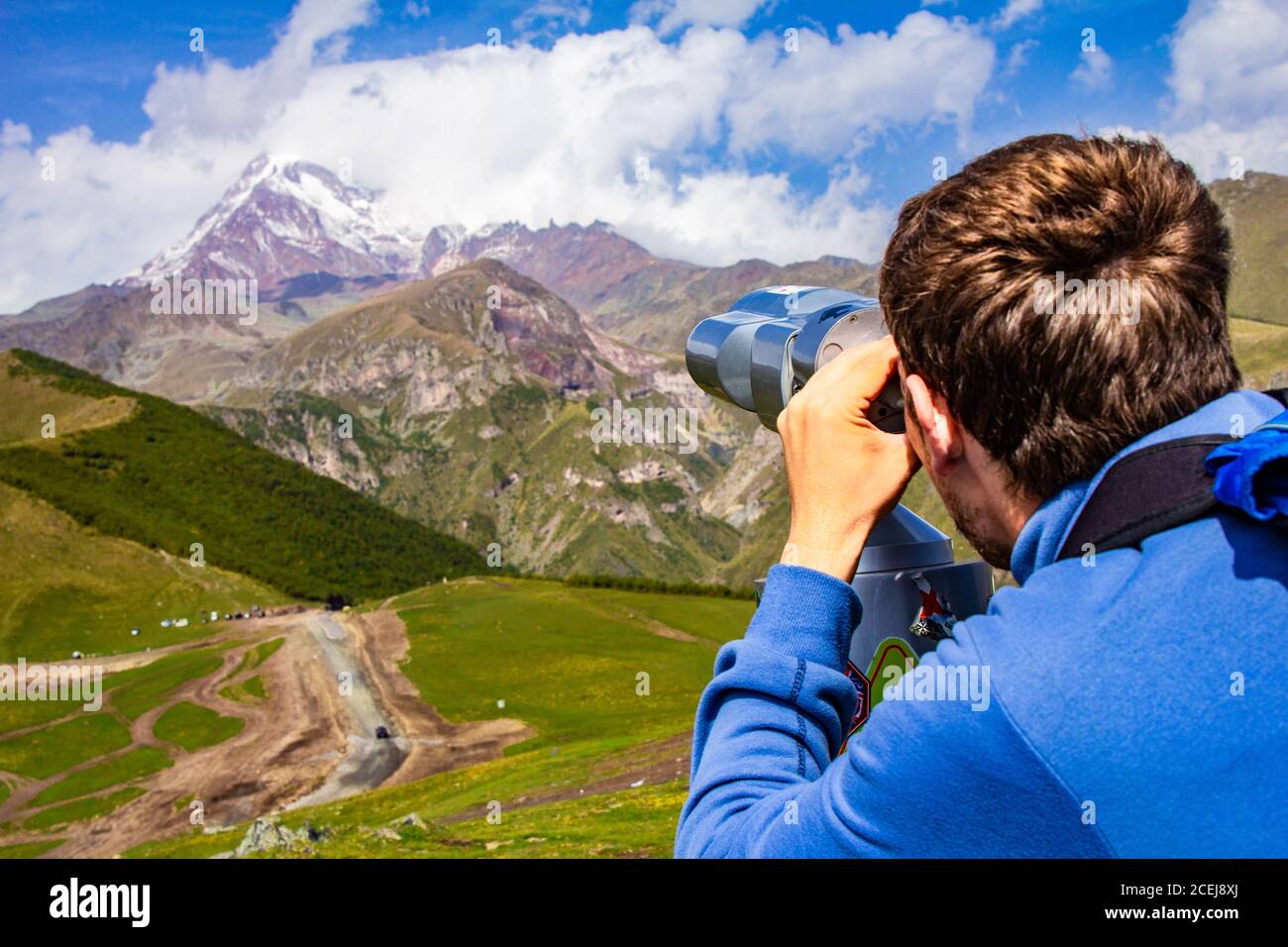 Mann, der im Binoskop auf die verschneiten Berggipfel gerichtet schaut Von Kazbeg aus der Sicht von Gergeti Trinity Church Tsminda Sameba Heilige Kirche in der Nähe Stockfoto