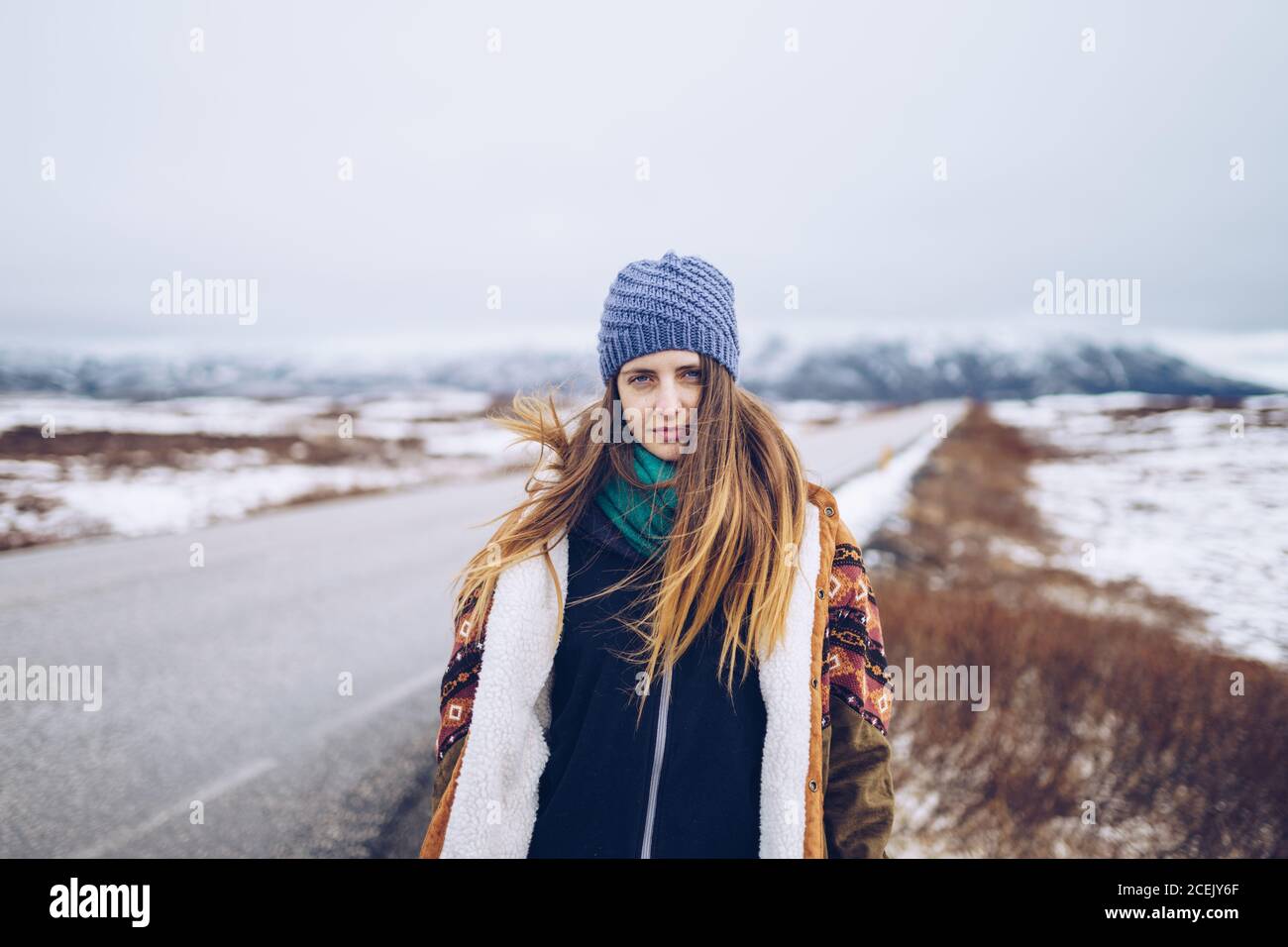 Junge attraktive Dame in Skijacke und Hut Blick auf Kamera auf der Straße zwischen wilden Ländern im Schnee in Island Stockfoto