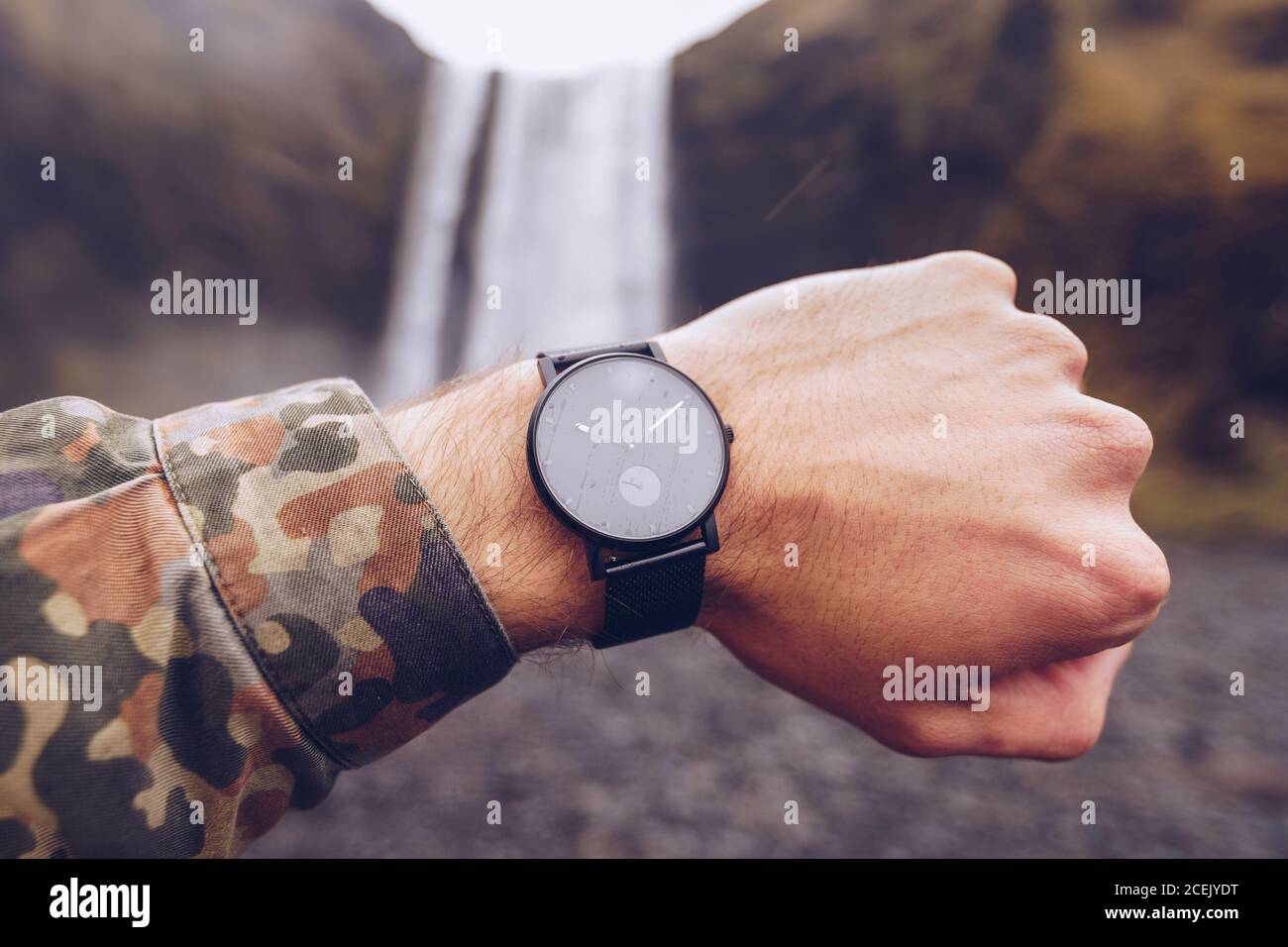 Crop Hand von Kerl zeigt schwarze Uhren in der Nähe von Wasser Kaskade In Island auf verschwommenem Hintergrund Stockfoto