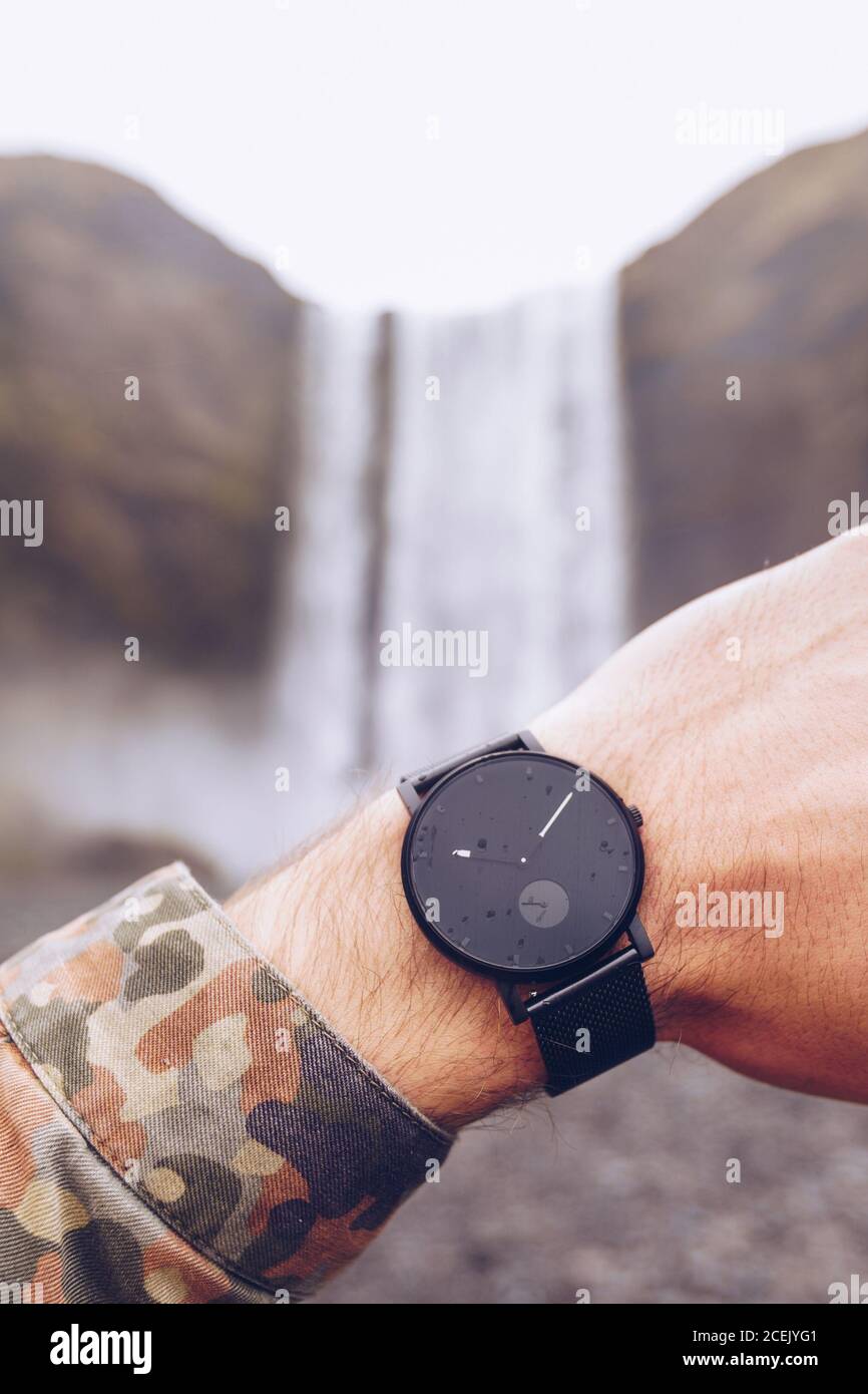 Crop Hand von Kerl zeigt schwarze Uhren in der Nähe von Wasser Kaskade In Island auf verschwommenem Hintergrund Stockfoto