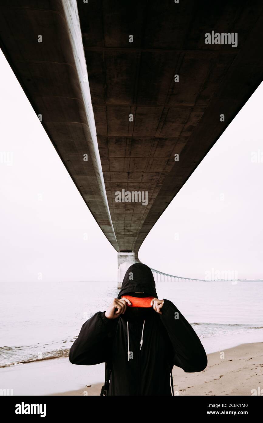 Guy in schwarz tragen halten Peak und stehen an der Küste Unter Brücke über Wasser und wolkenbewölkter Himmel in Frankreich Stockfoto