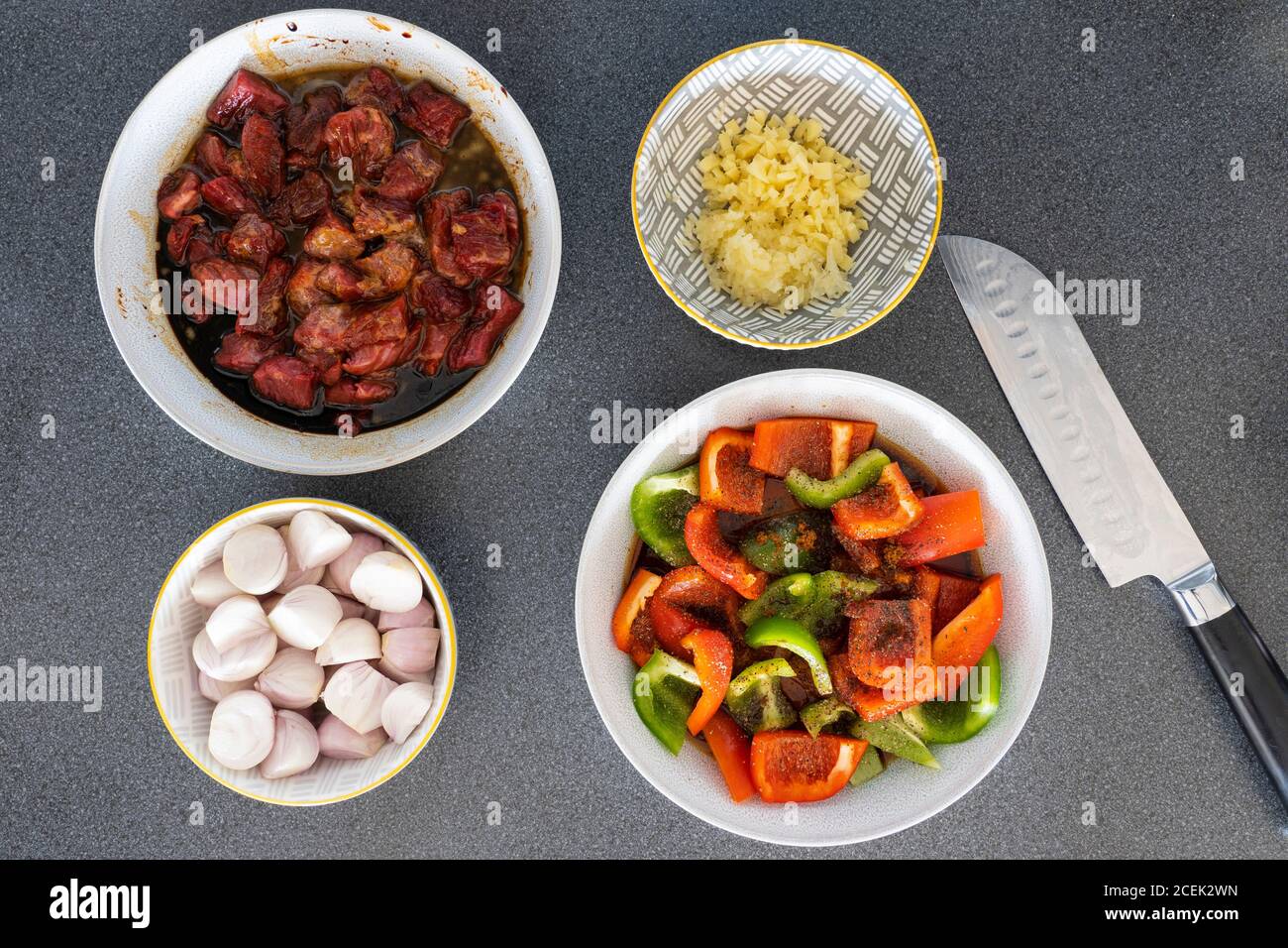 Vorbereitete gehackte Rohzutaten - Rindfleisch, Paprika, Zwiebeln, Knoblauch und Ingwer in Schüsseln mit einem japanischen Messer bereit für das Kochen einer Rührfritte Gericht Stockfoto
