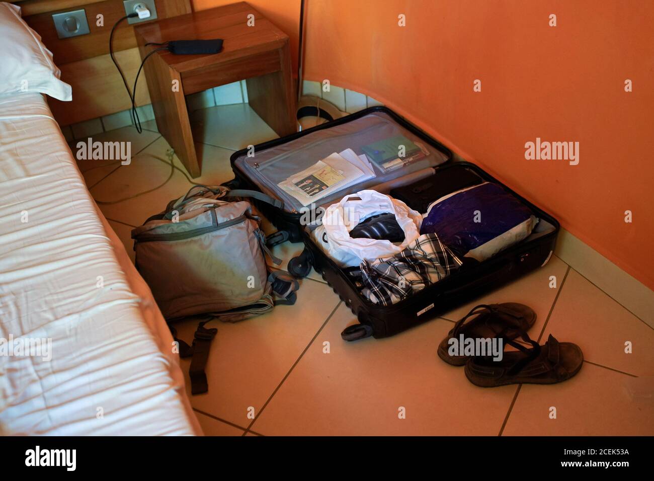 Unordentlicher schwarzer Gepäckkoffer, der auf dem Boden des Hotelzimmers geöffnet wurde, Rucksack und ein Paar Sandalen daneben. Stockfoto