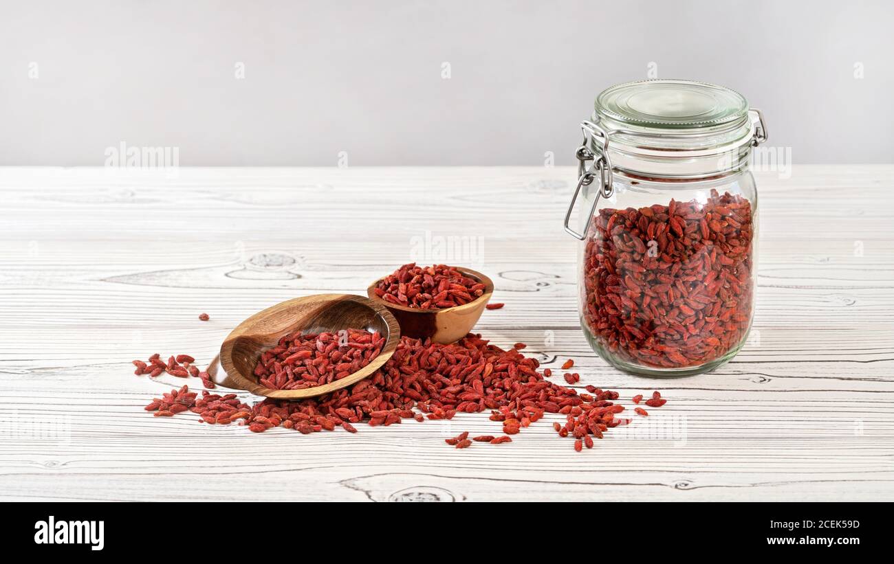 Getrocknete Goji aka. wolfberry innen Glasflasche, einige in Holzschüsseln und verschüttet auf weißen Brettern Schreibtisch, Platz für Text links oben Stockfoto