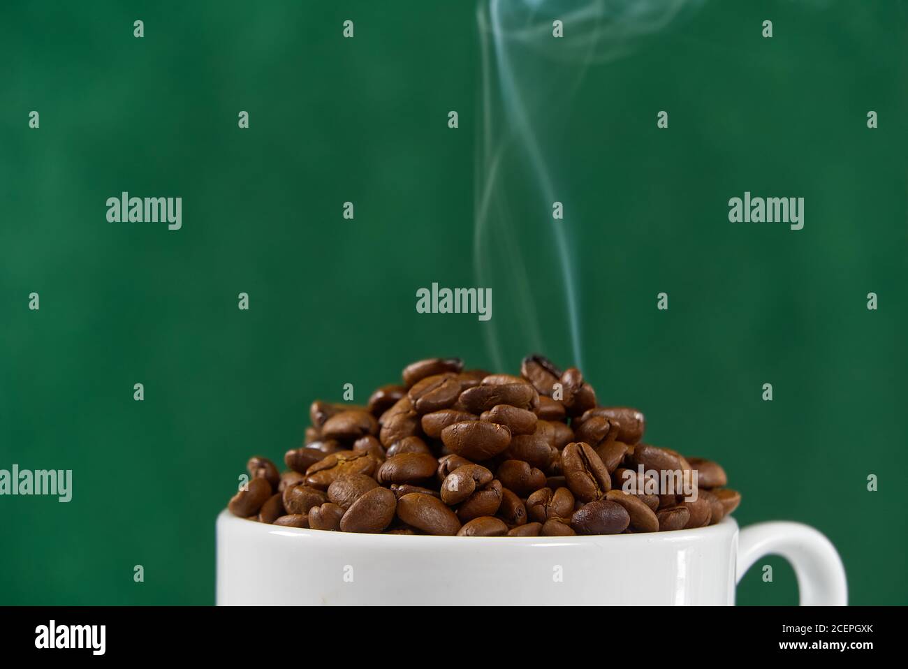 Internationaler Tag des Kaffees Konzept. Nahaufnahme weiße Kaffeetasse voller Kaffeebohnen auf grünem Hintergrund mit Rauch auf der Oberseite. Stockfoto