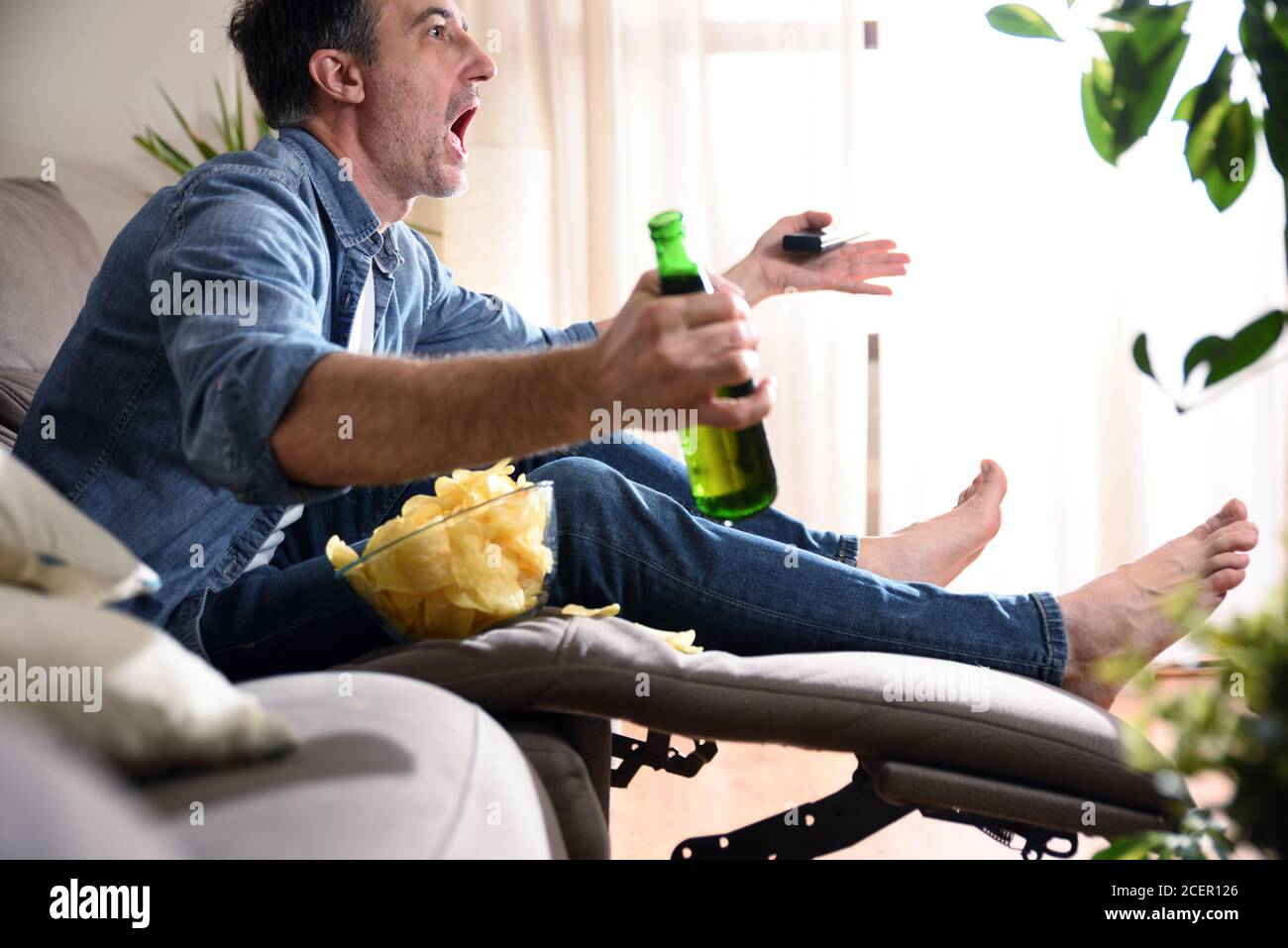 Wütender Mann, der Sport im Fernsehen sieht, sitzt bequem auf einem Sofa und hat einen Snack mit Bier und einer Fernbedienung in den Händen. Stockfoto