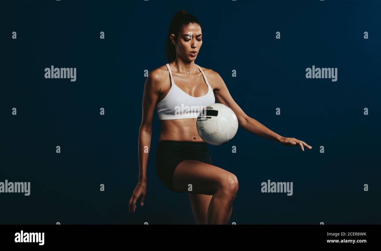 Frau Fußballspielerin konzentriert sich auf Jongliertechniken. Weiblich in Sportkleidung jongliert einen Ball mit auf dem Knie über dunklem Hintergrund. Stockfoto