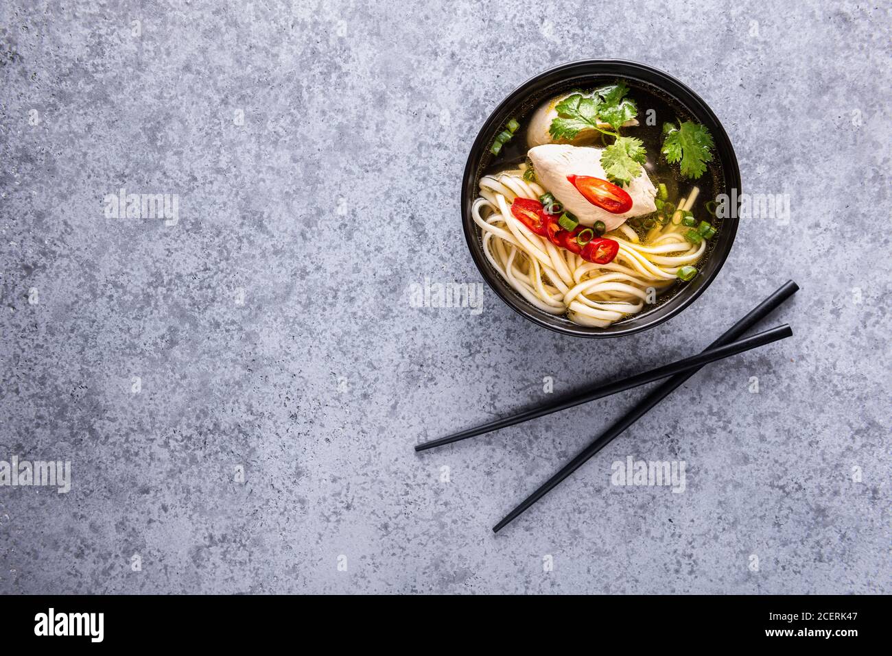 Würzige asiatische Suppe. Hühnerbouillon und Fleisch, rote Chilischote, Koriander, Frühlingszwiebeln und japanische Nudeln in schwarzer Schüssel mit Essstäbchen auf grauer Oberfläche Stockfoto